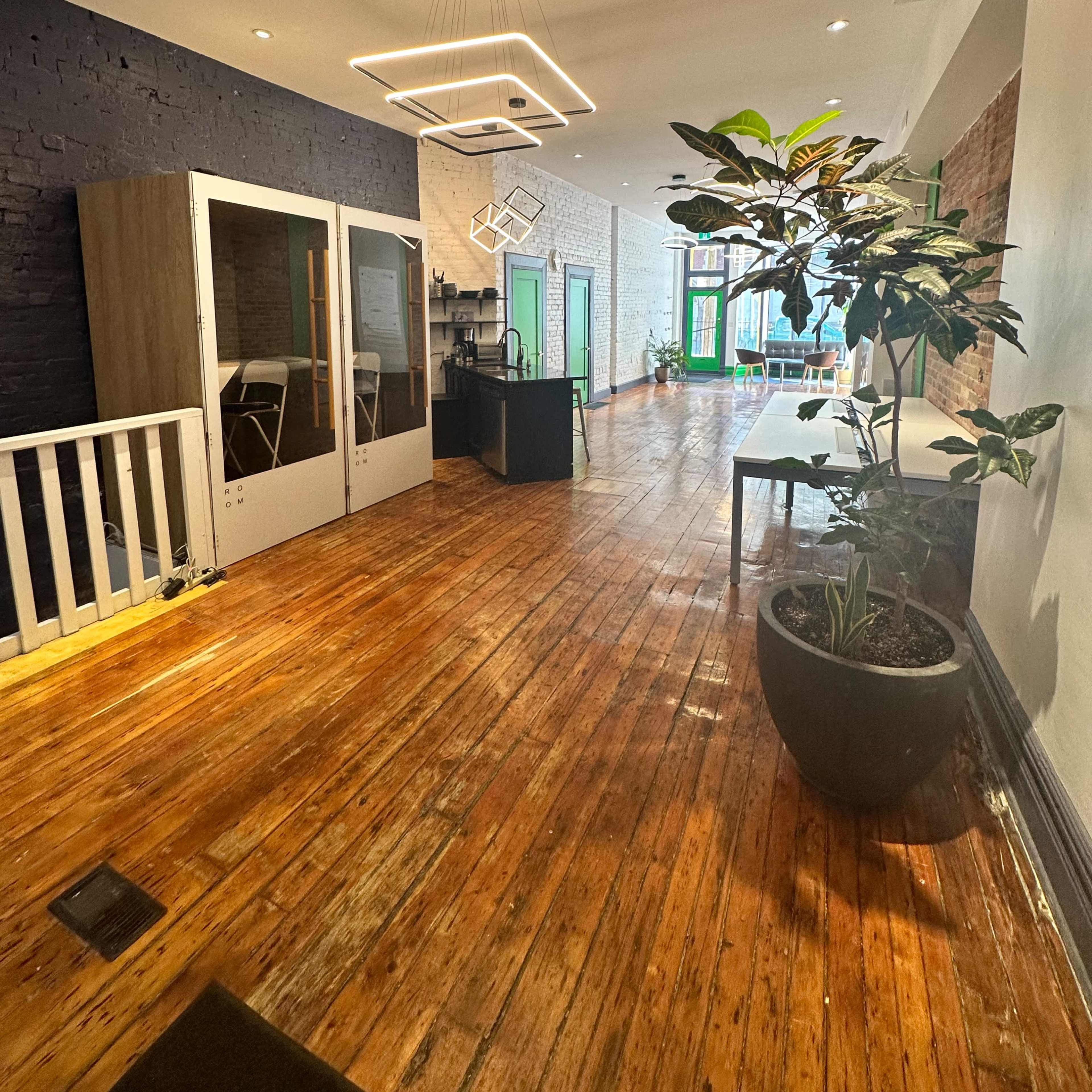 A modern office interior with wooden floors, large windows, and a mix of open and enclosed workspaces.