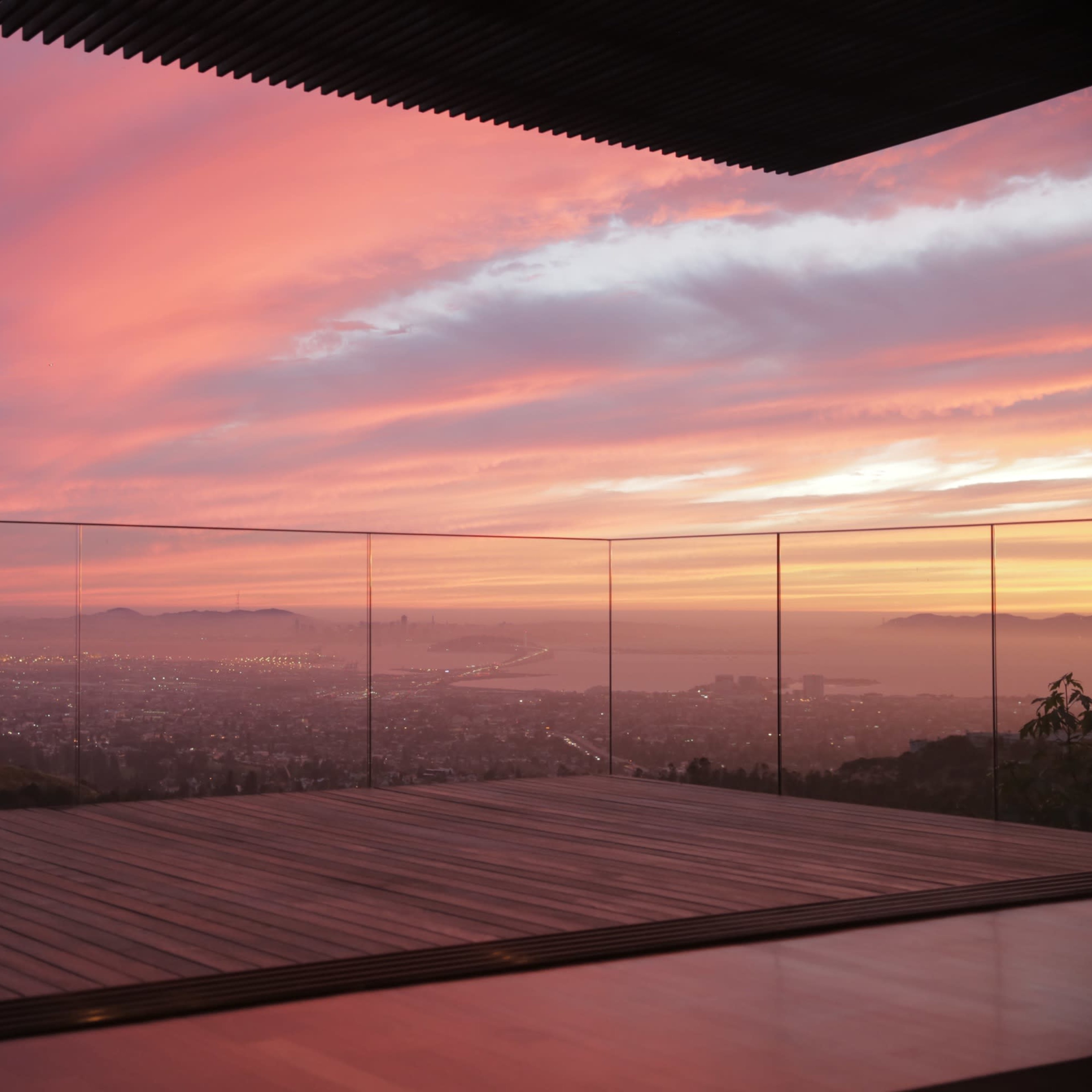 A glass-enclosed balcony overlooking a city skyline at sunset, with vibrant clouds reflecting shades of pink and orange in the sky.