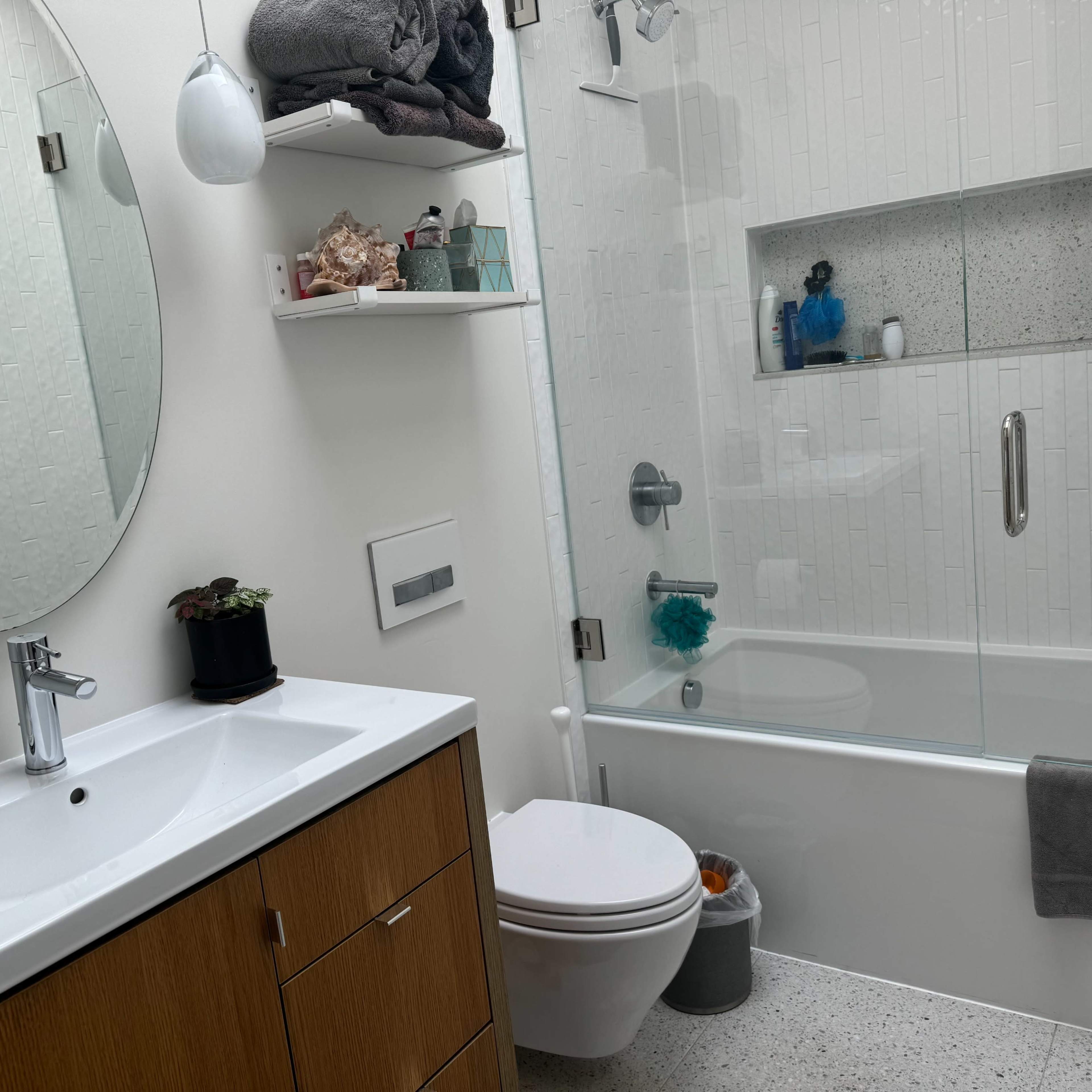 A modern bathroom features a glass shower, white tiled walls, a circular mirror, and a wooden vanity with a sink.
