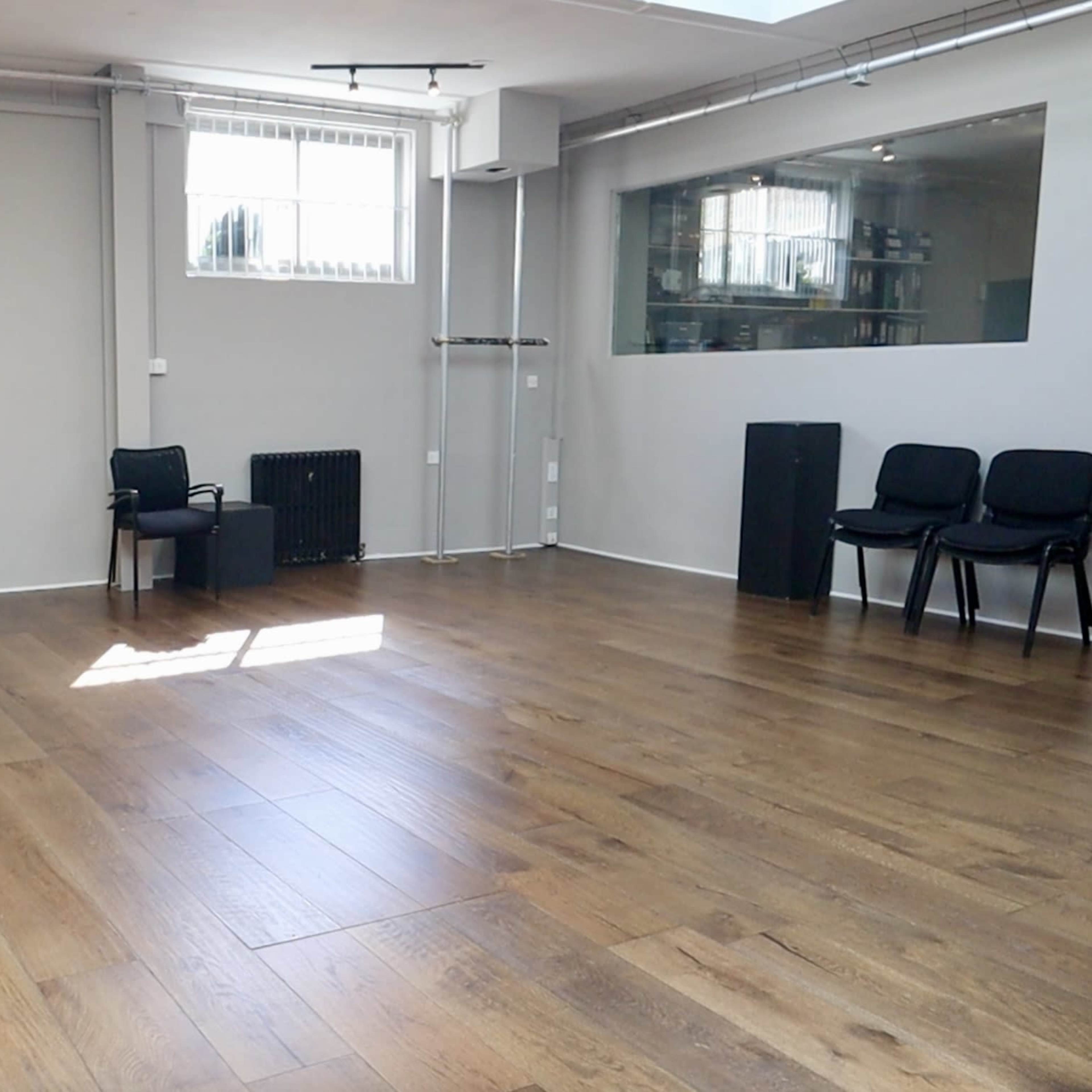 An empty, well-lit room with wooden flooring, a large window, and a few black chairs arranged in the space.