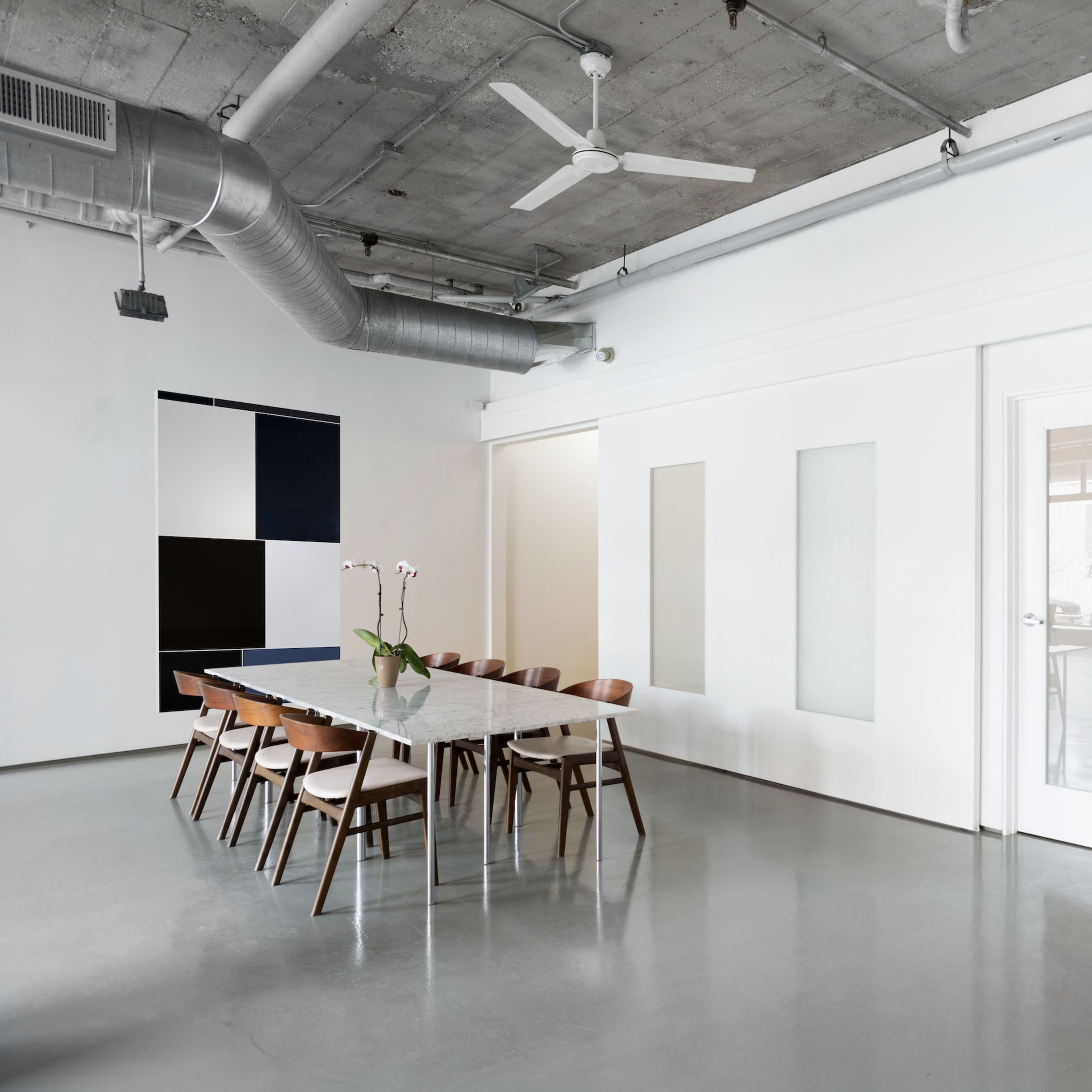 A modern, minimalist interior with a long dining table surrounded by chairs, positioned in a spacious room with white walls and concrete flooring.