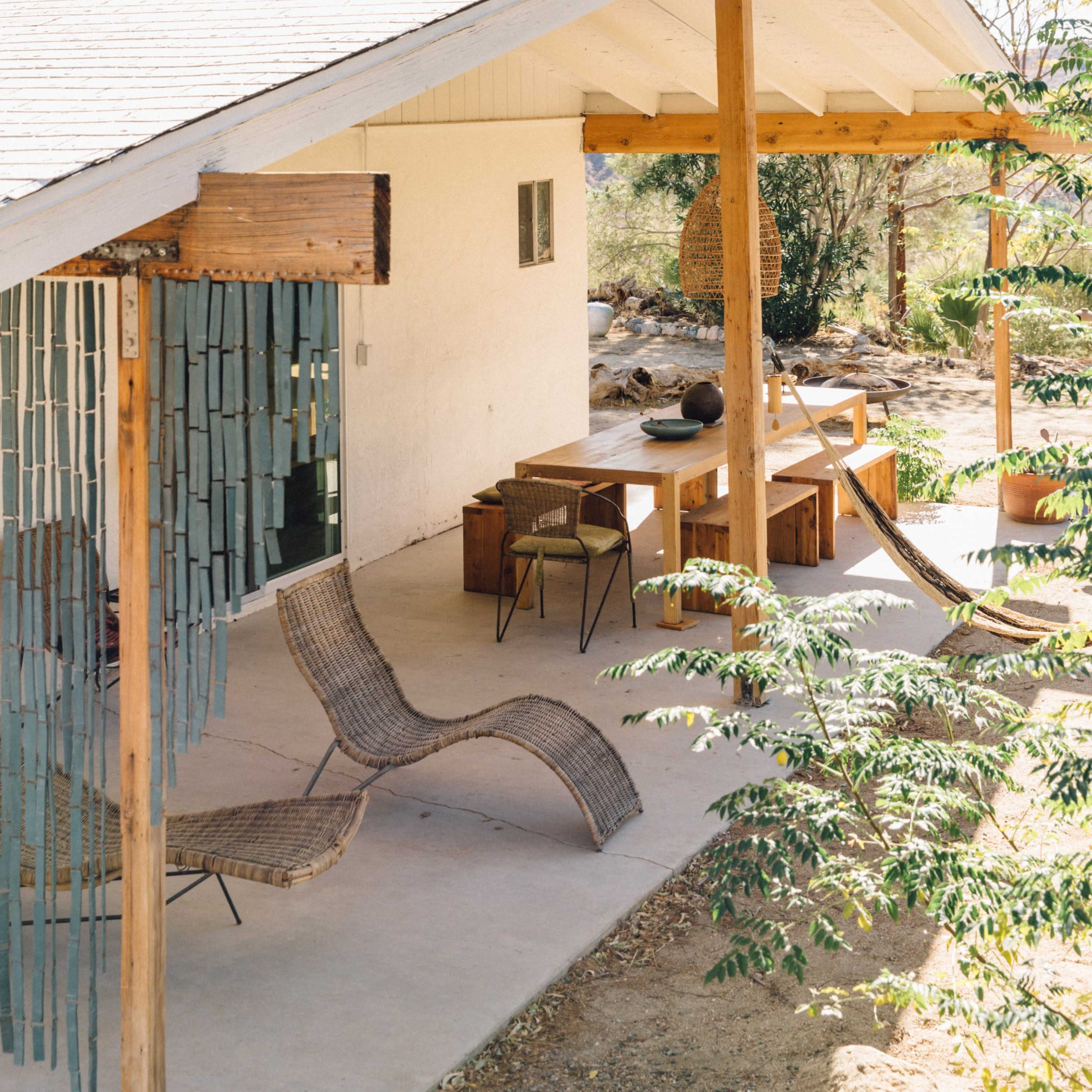 A modern house features a spacious porch with wooden posts, woven chairs, and a dining table surrounded by trees.