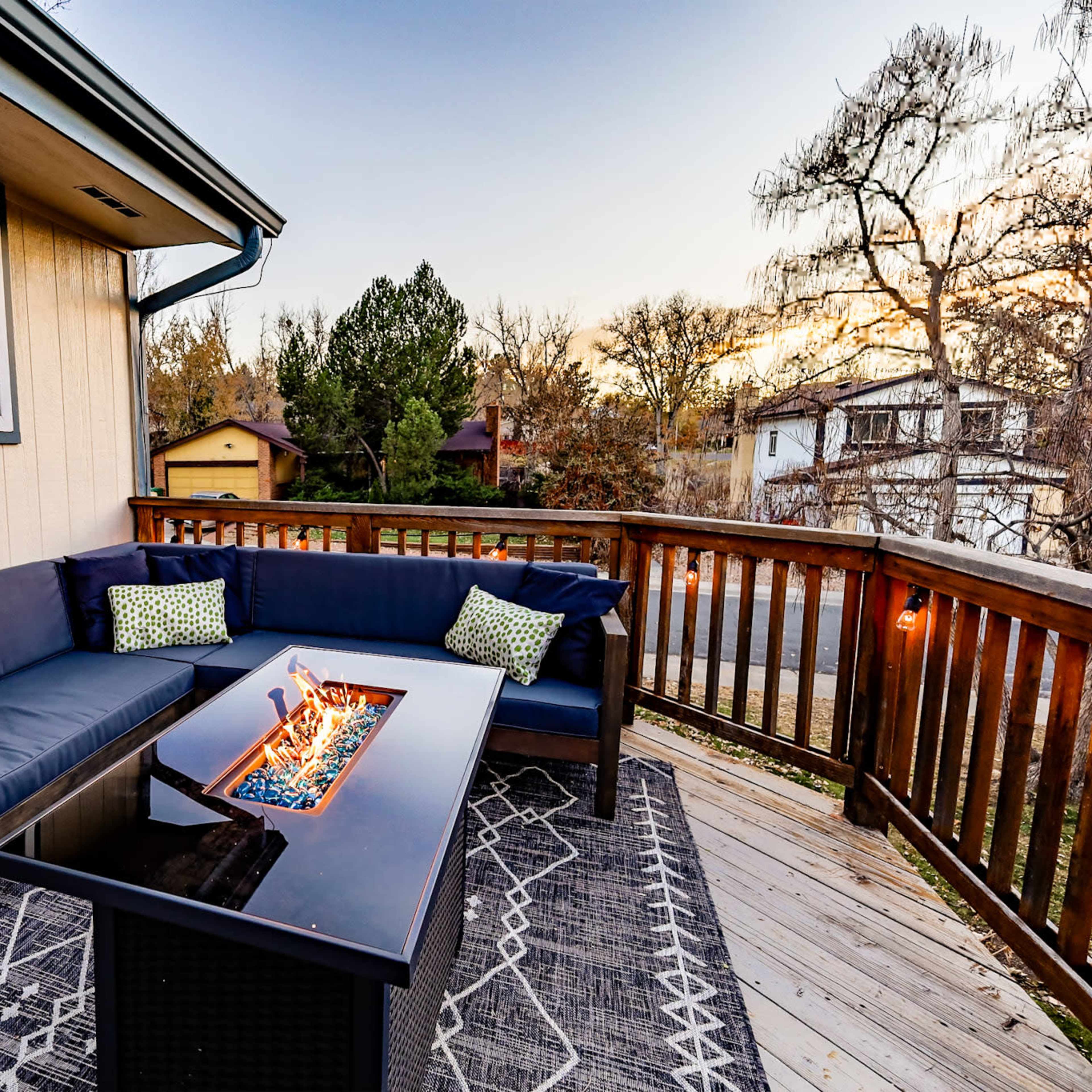 A deck features a sectional sofa with decorative cushions surrounding a fire pit on a wooden surface.