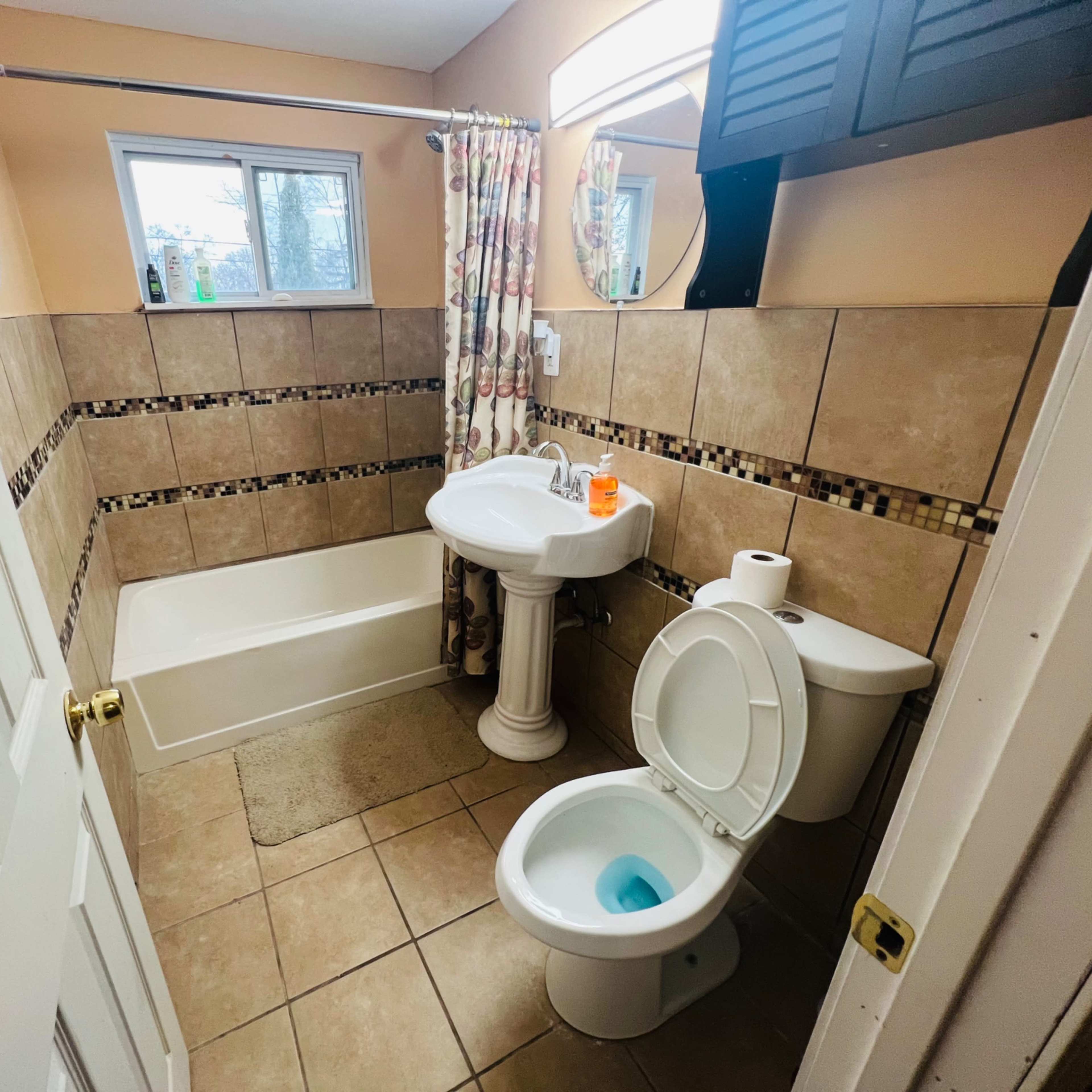 A bathroom with a bathtub, pedestal sink, and toilet, featuring tiled walls and a window.