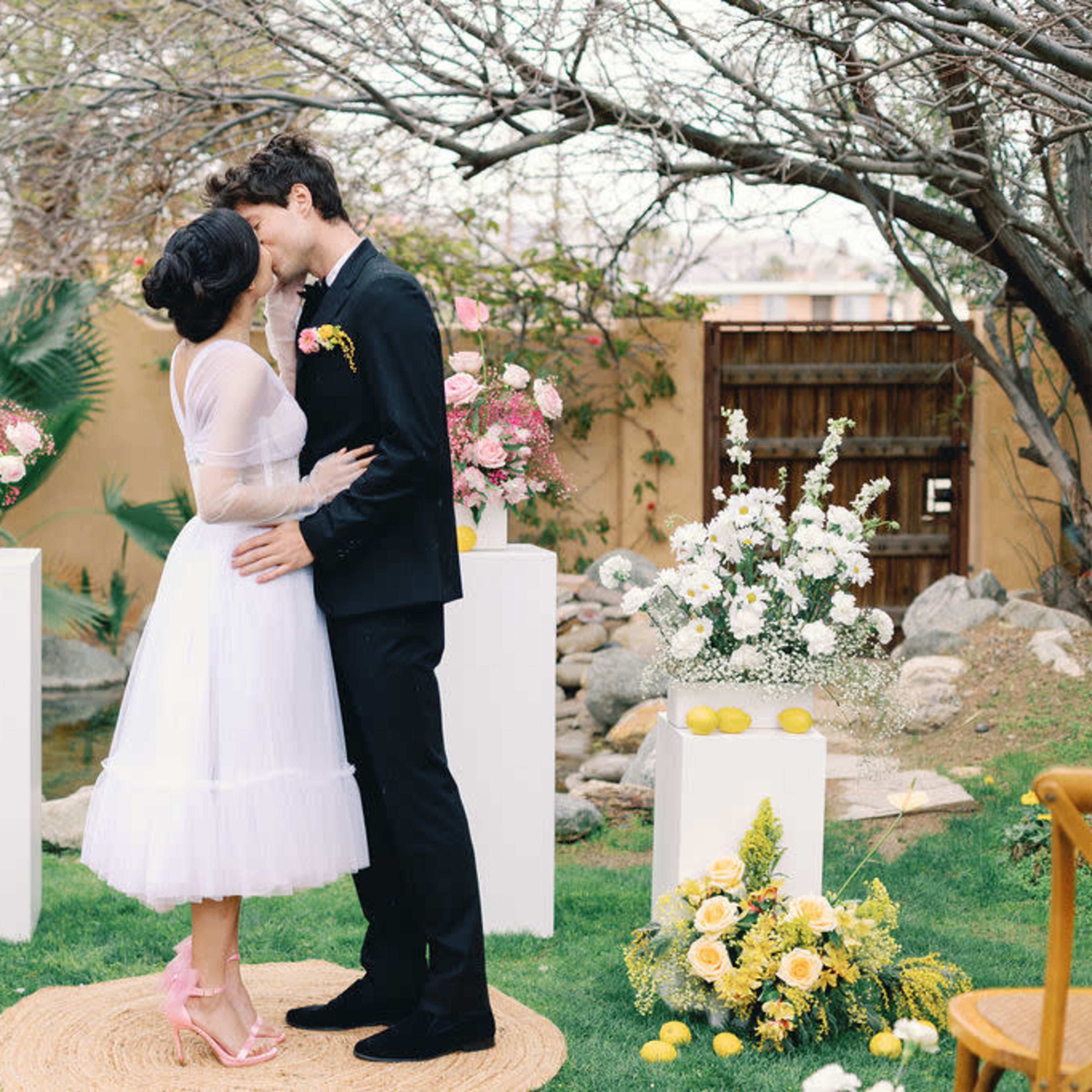 A couple kisses in front of a floral wedding altar adorned with white flowers and greenery in a garden setting.