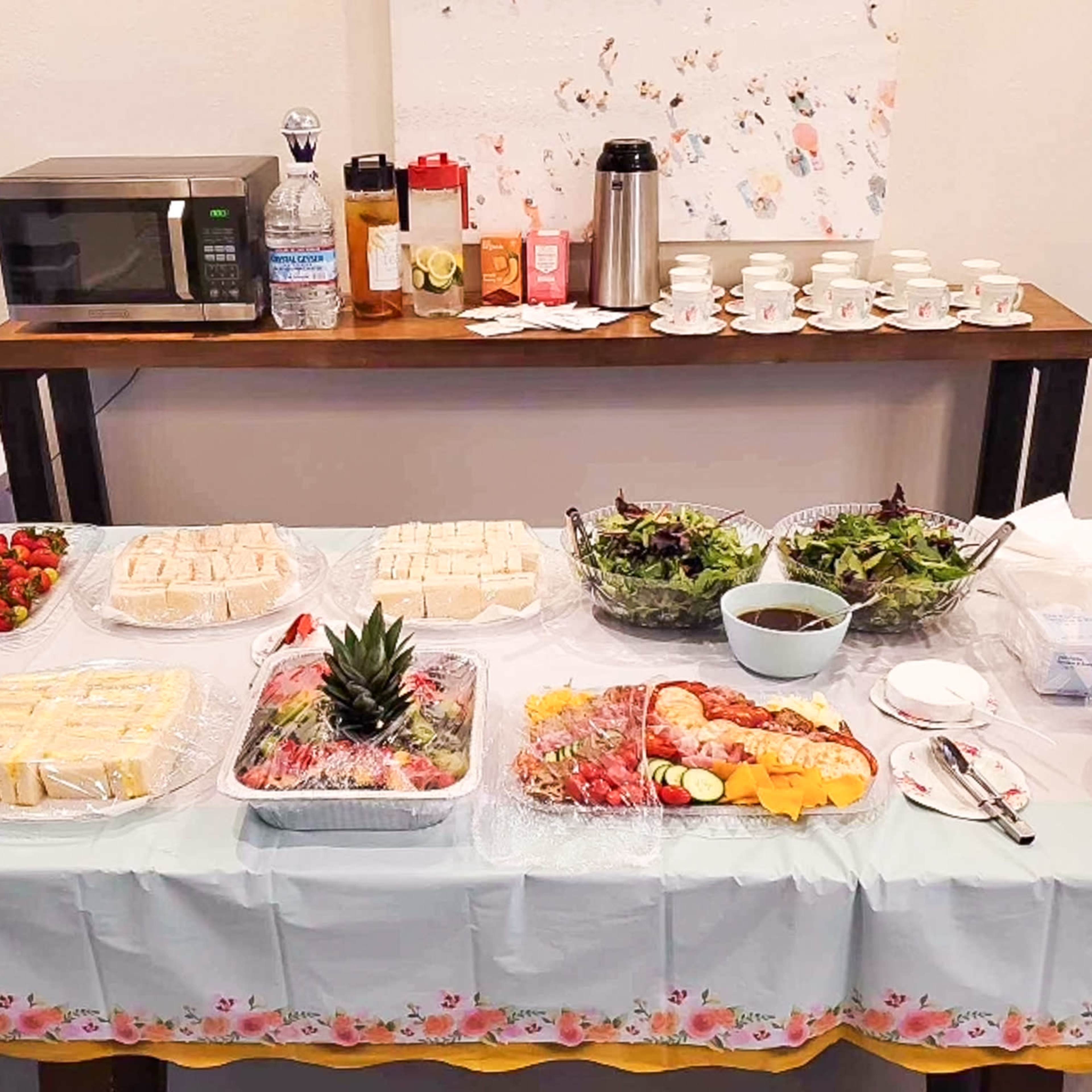 A buffet table is set with various food items, including sandwiches, salads, fruit platters, and beverages, against a wall with a mounted drawing.