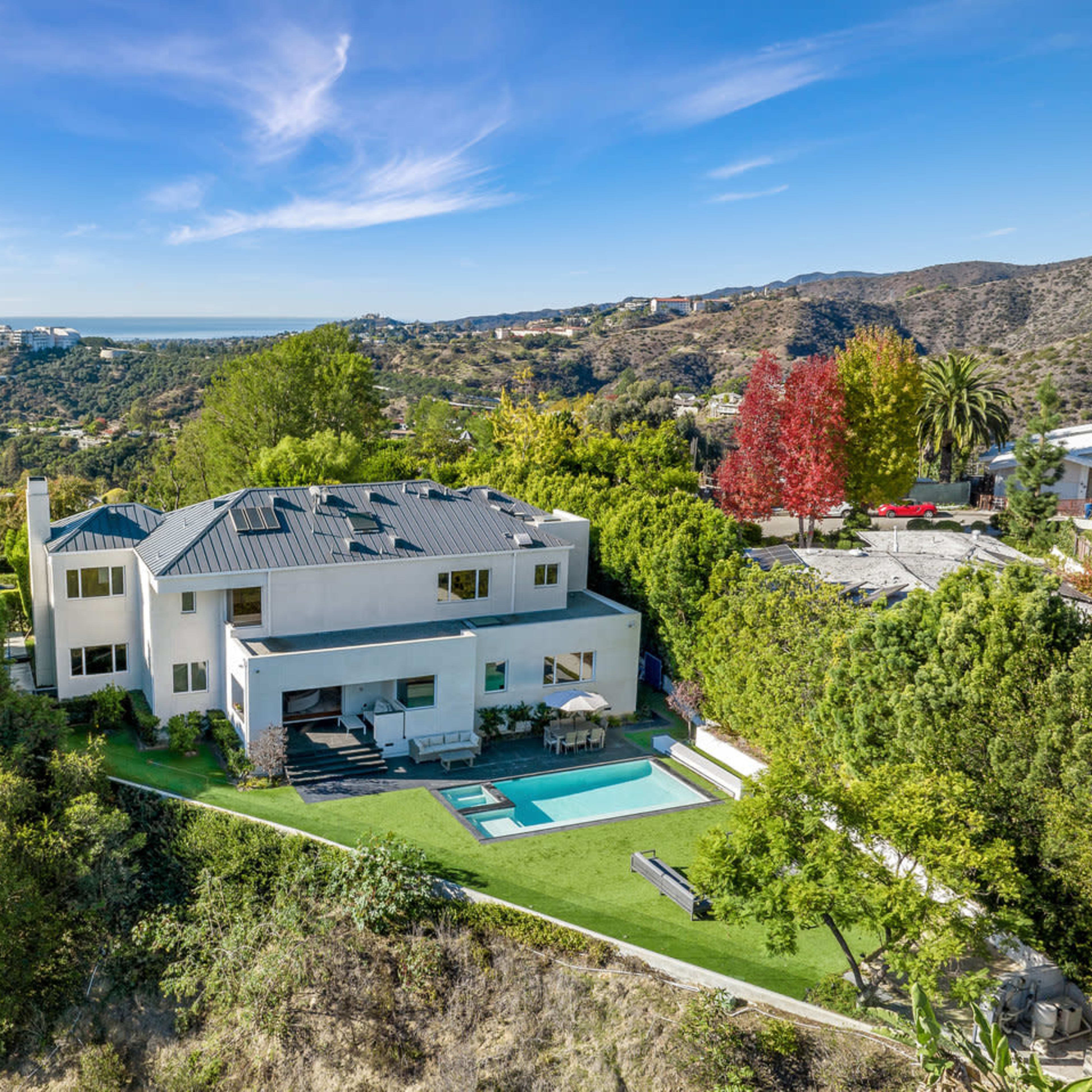 A modern two-story house with a pool is set on a hillside, surrounded by greenery and featuring a view of the distant hills.