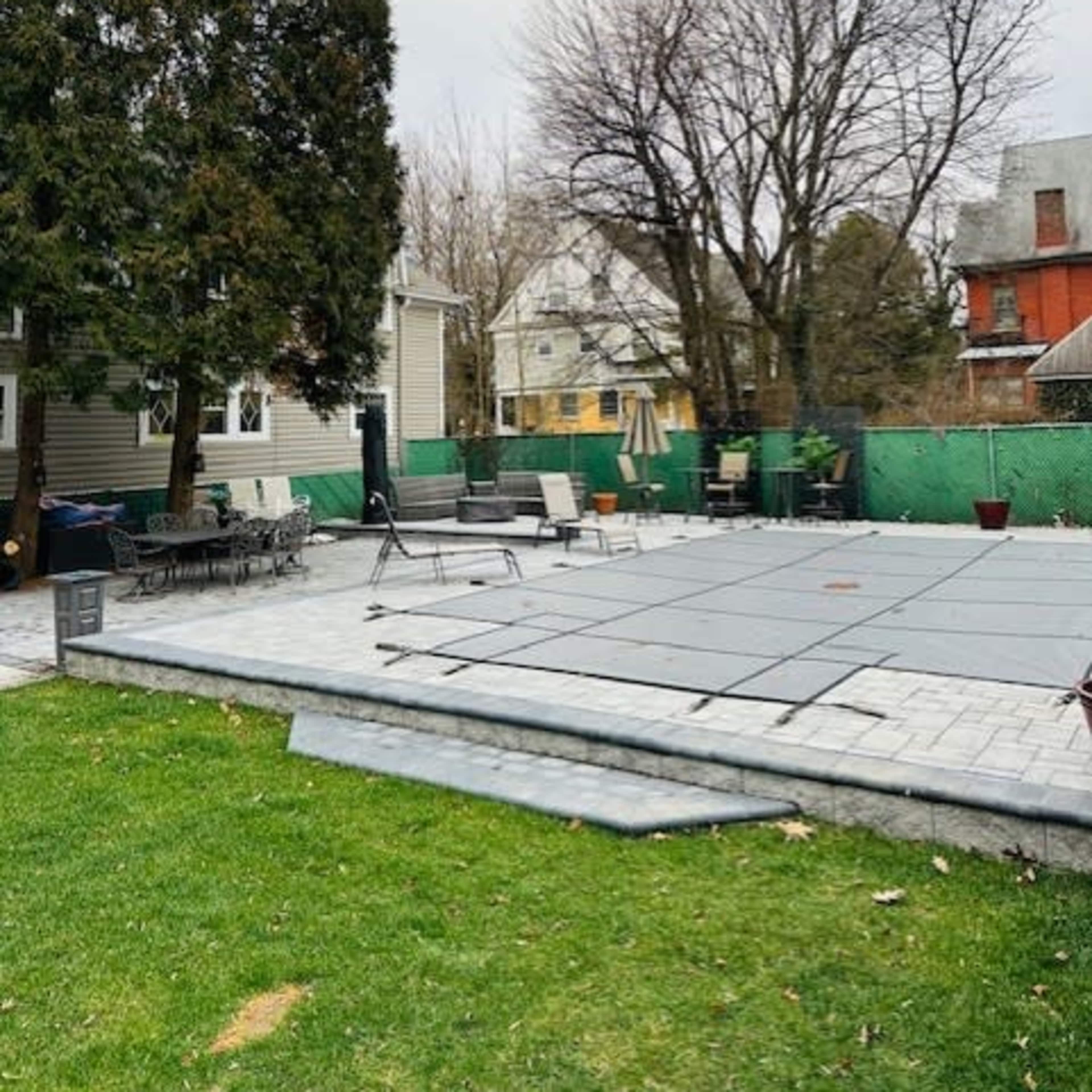 A backyard scene features a swimming pool area covered with a safety cover, surrounded by patio furniture and trees.
