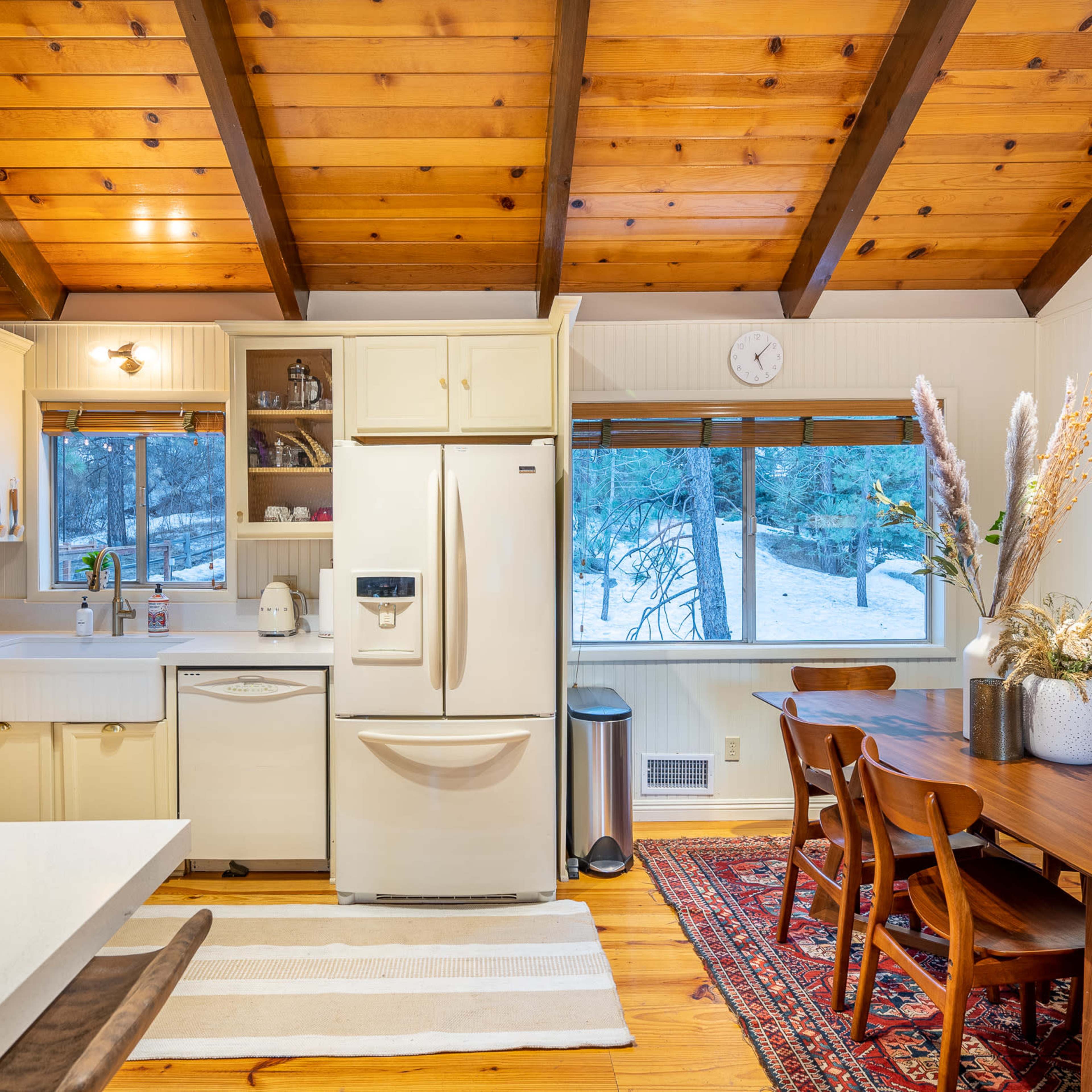 A kitchen with wooden beams, light-colored cabinets, and a dining table, featuring a view of a snowy outdoor scene through a window.