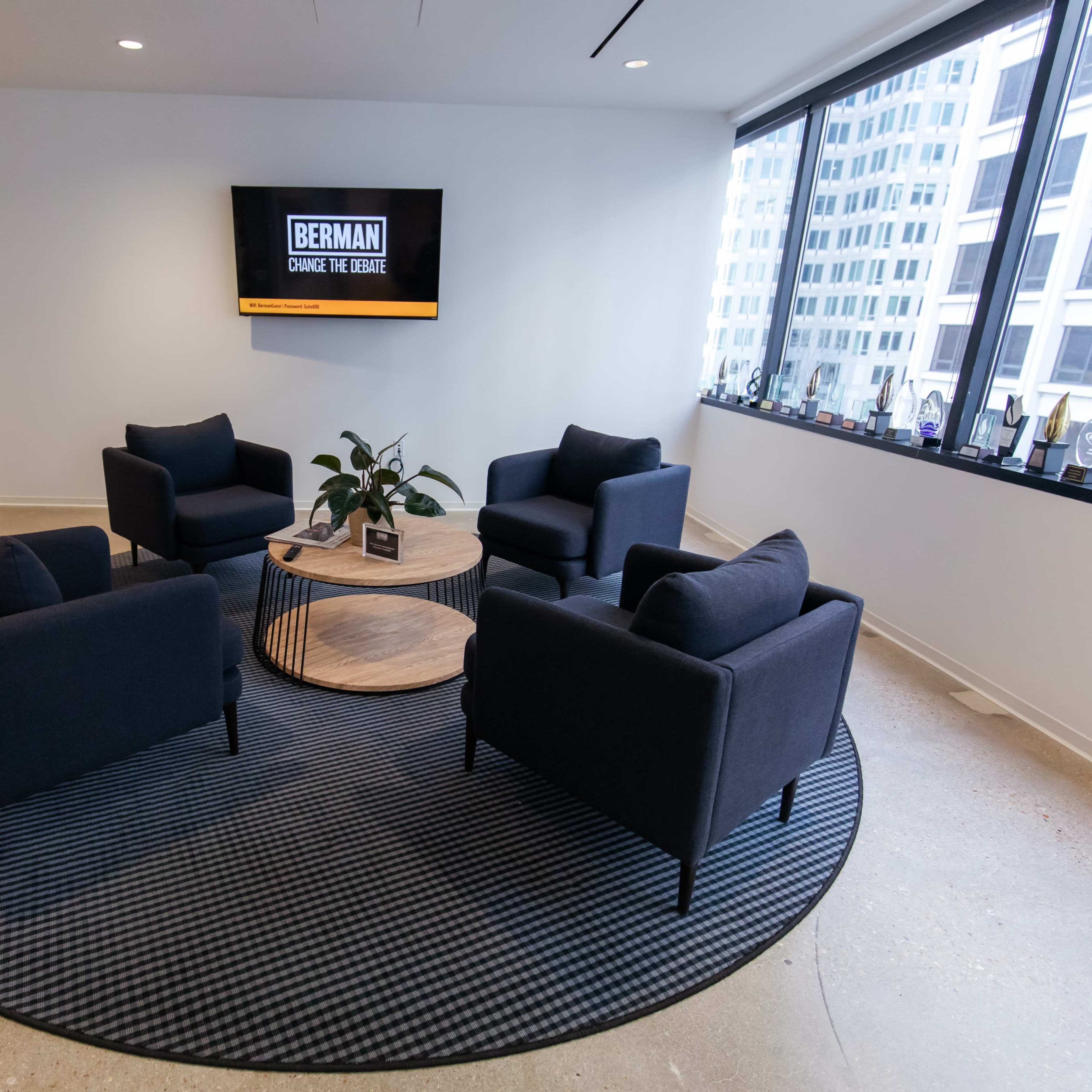 A modern office lounge area features four black chairs arranged around a round wooden coffee table on a patterned rug, with a television mounted on the wall displaying the name "BERMAN."