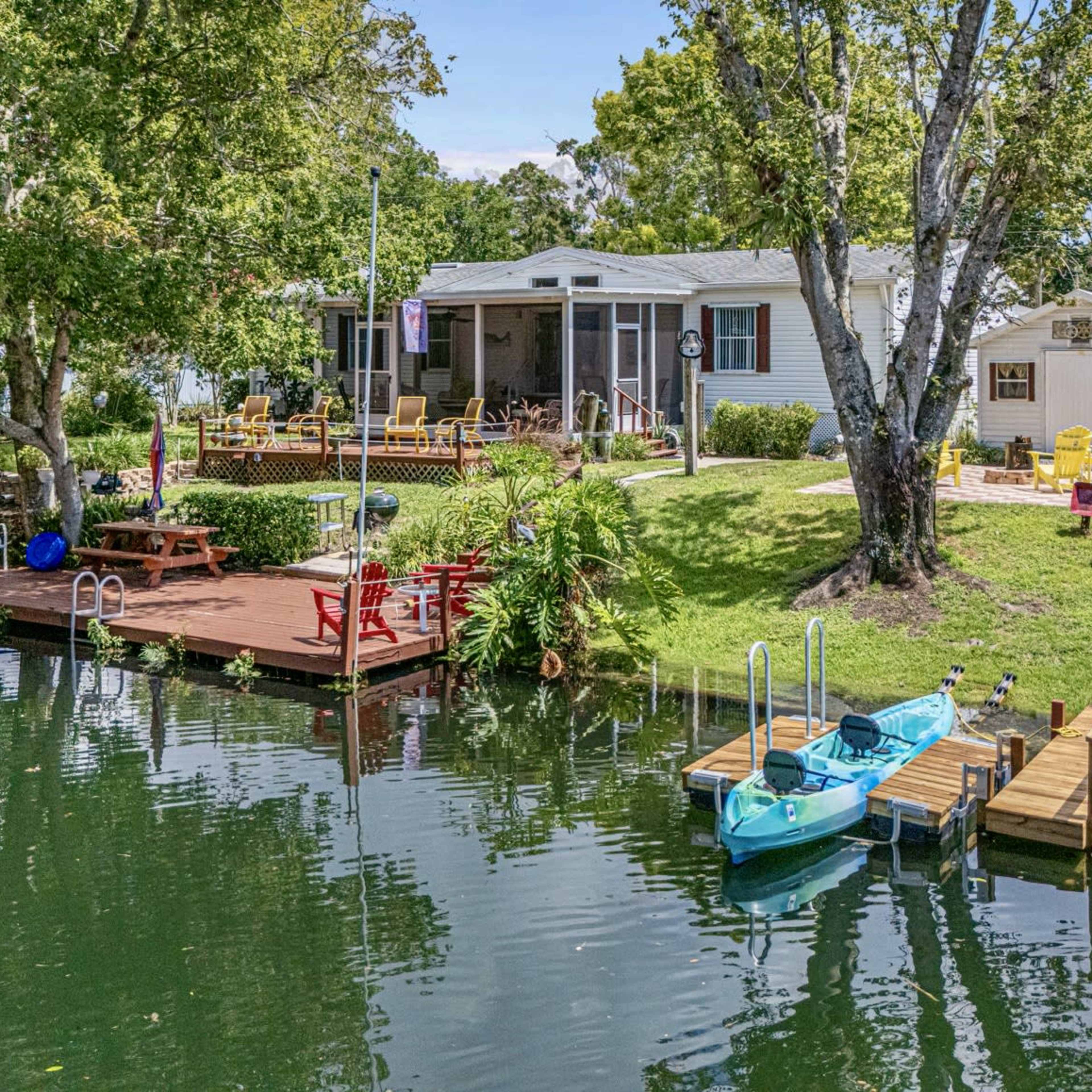 A waterfront property features a wooden deck with seating, two kayaks, and lush greenery surrounding a home, all adjacent to calm water.