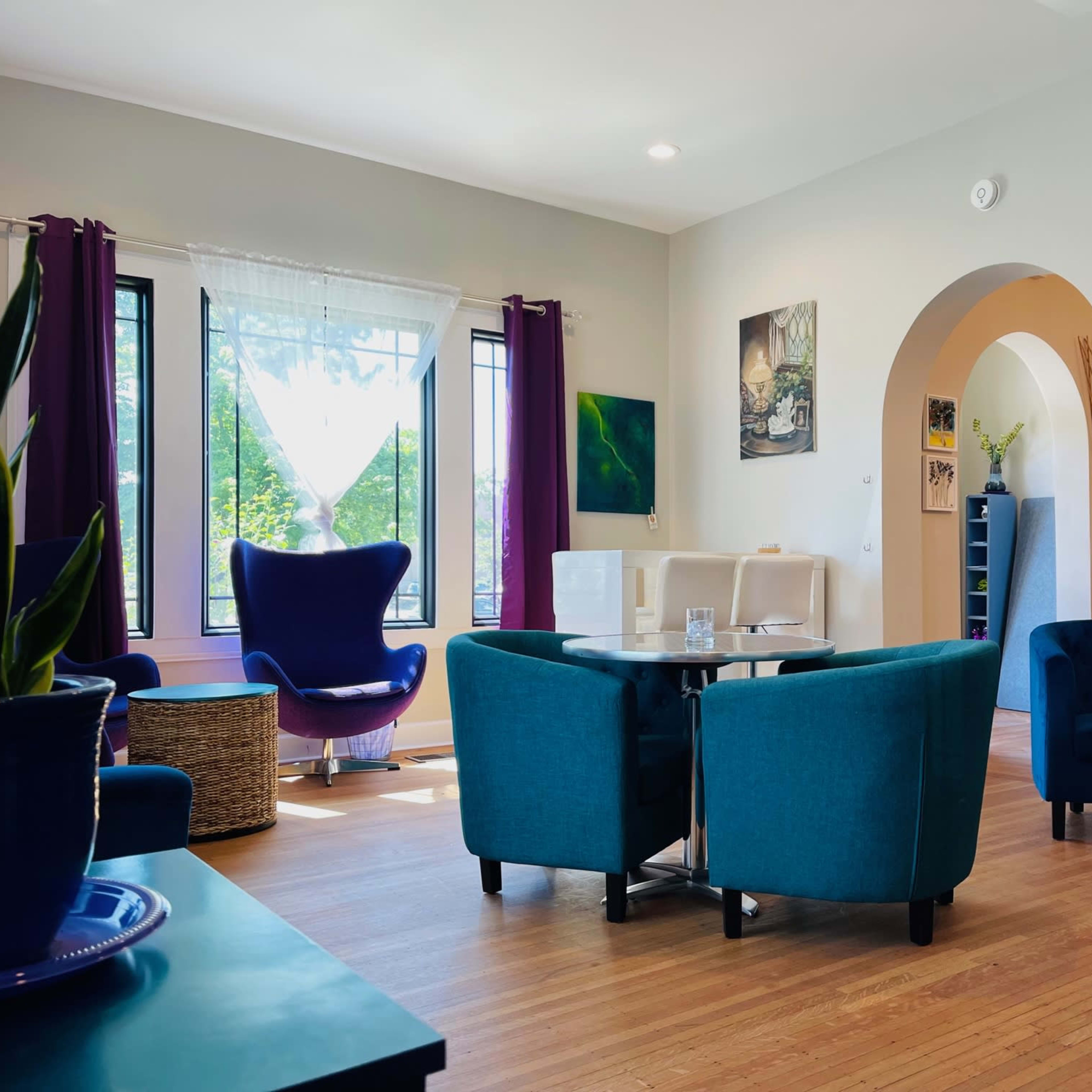 A modern, cozy lounge area with teal chairs and a large purple armchair, featuring large windows and light-colored walls.