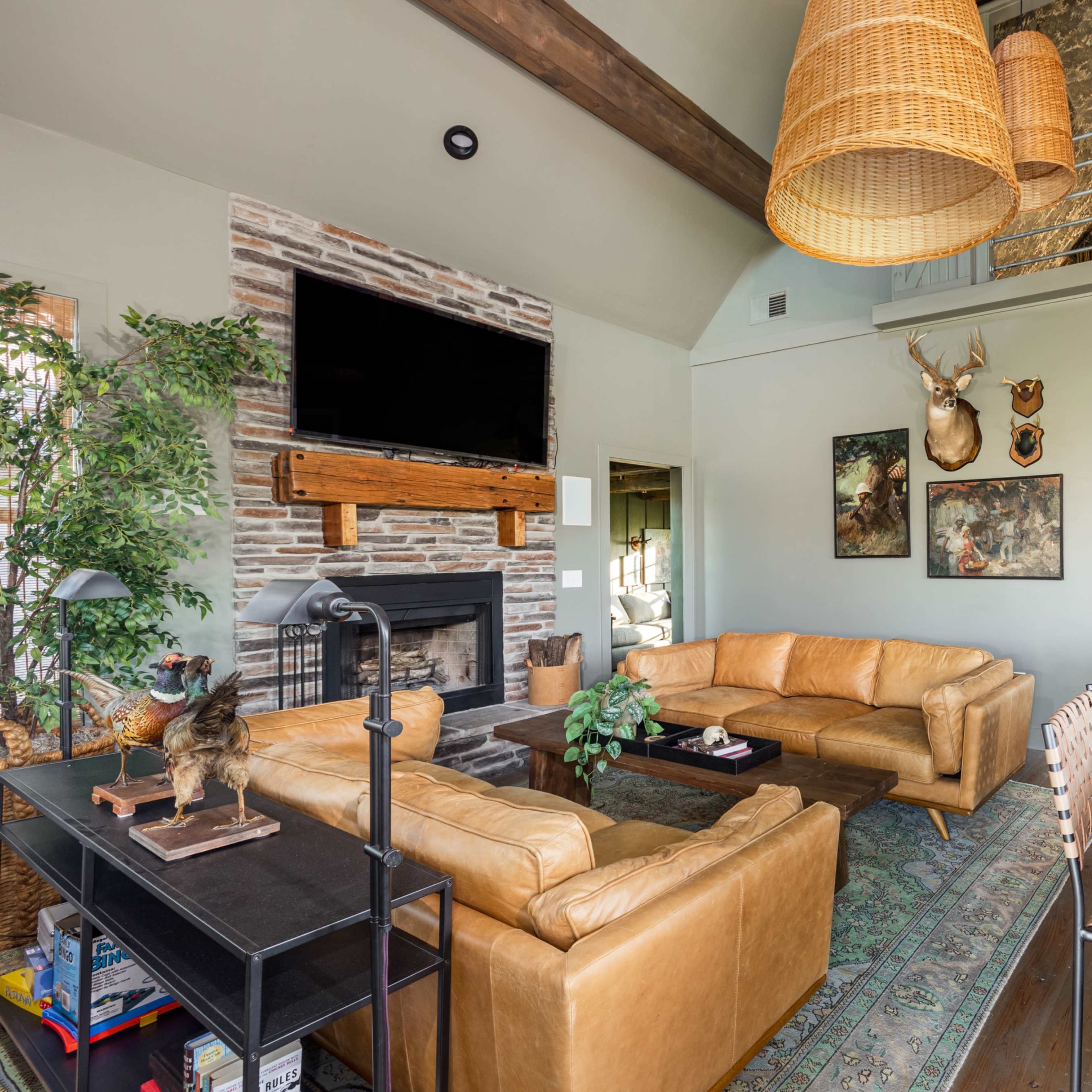 The living room features a leather sectional sofa, a stone fireplace, and a wooden dining table with metal chairs, surrounded by greenery and decorative artwork.