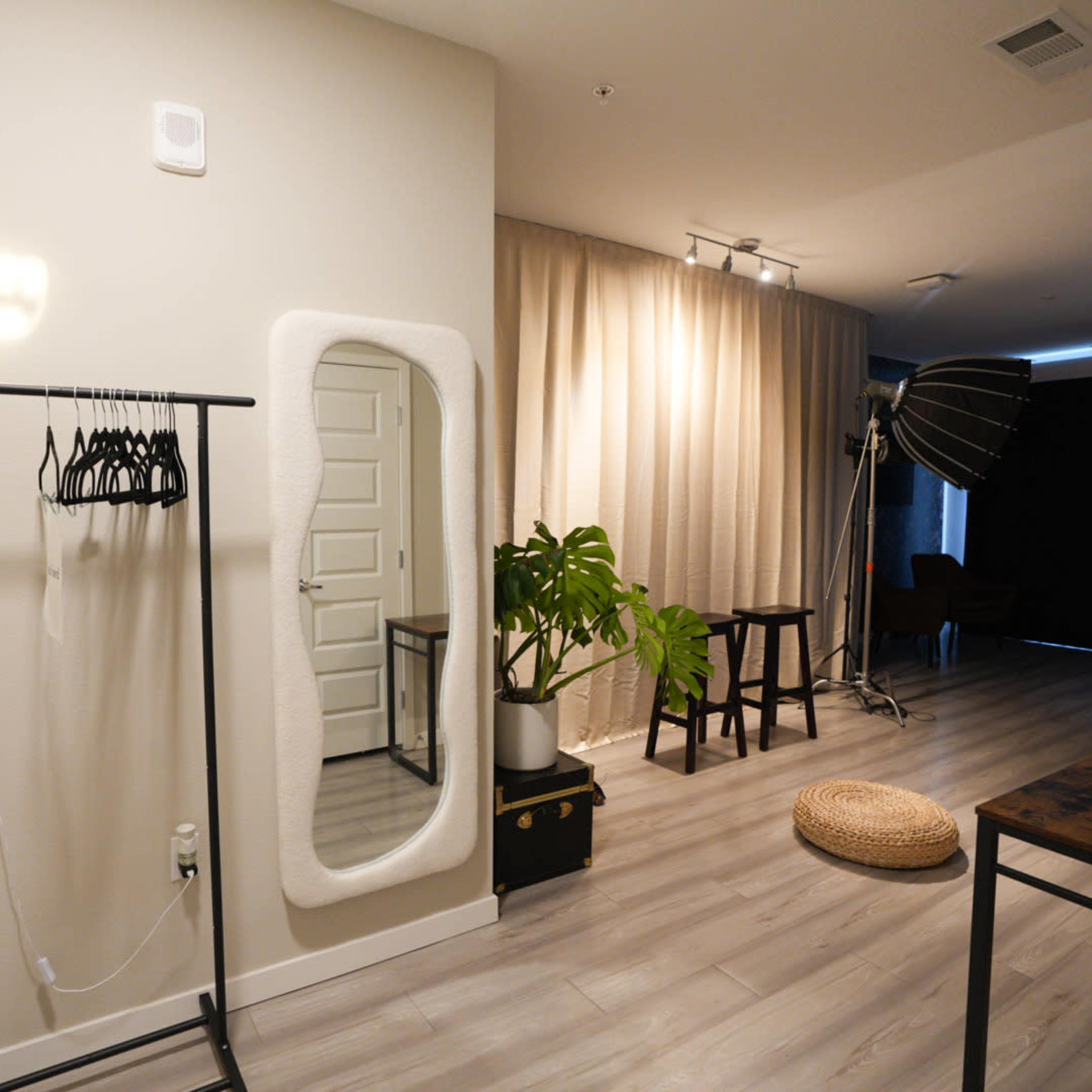 A minimalist interior space features a clothing rack, a large mirror, a potted plant, and a studio light setup against a neutral backdrop.