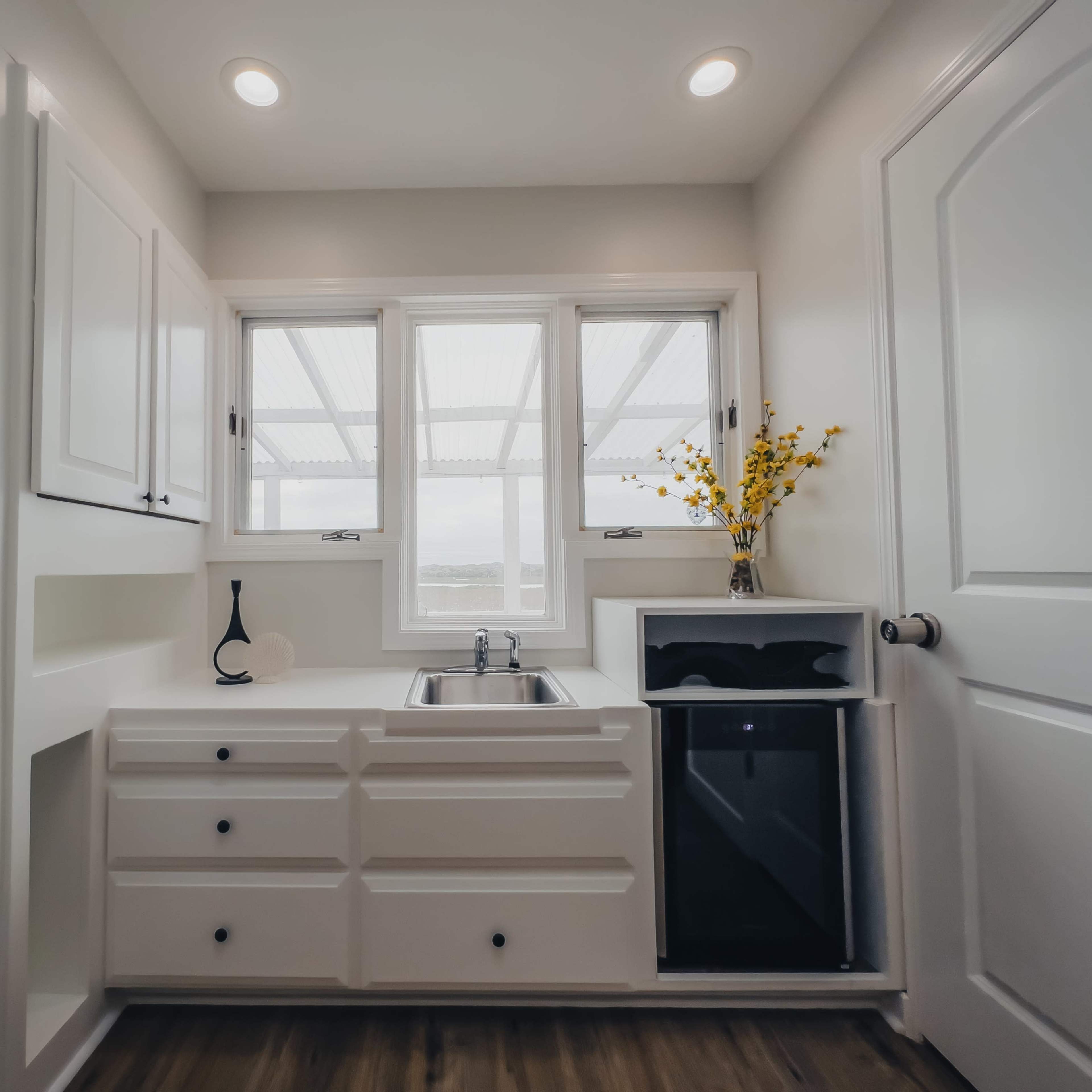 A small, modern kitchen area with a sink, a mini refrigerator, and large windows, featuring white cabinetry and a minimalist design.