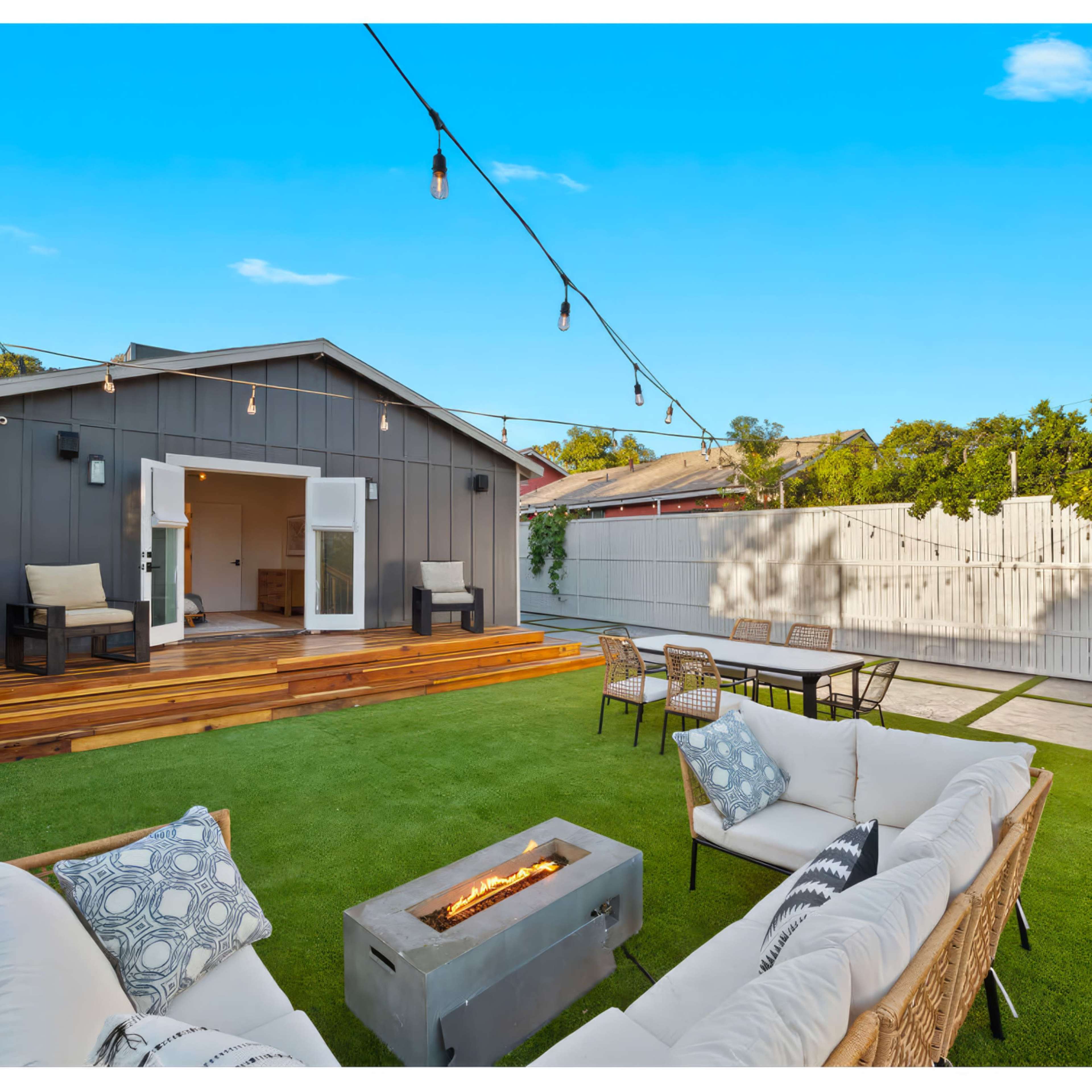A backyard features a modern gray house with a wooden deck, outdoor seating, a fire pit, and string lights overhead.