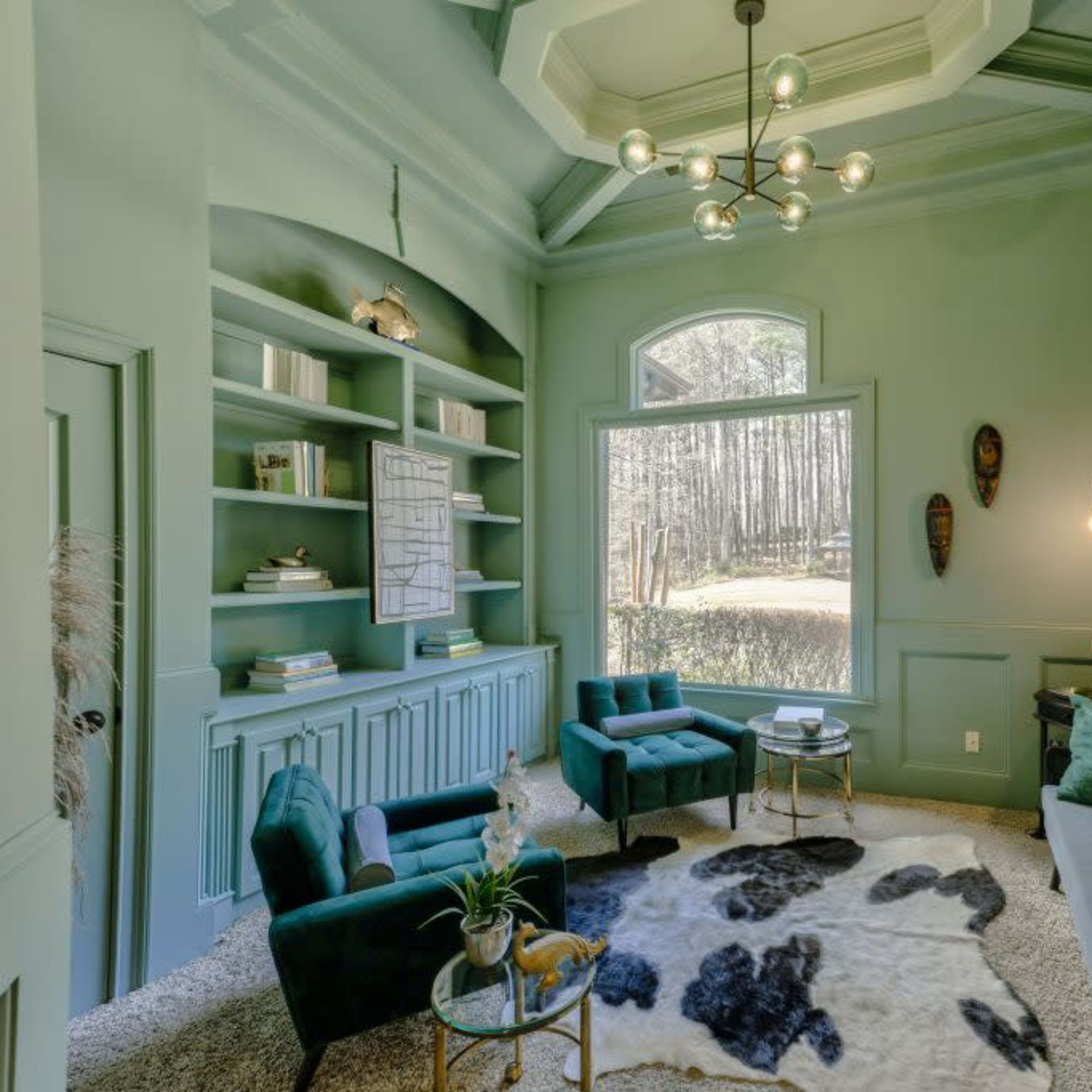 A small, light green room with a large window overlooking a wooded area, furnished with a sofa, two green chairs, a round table, and decorative accents.