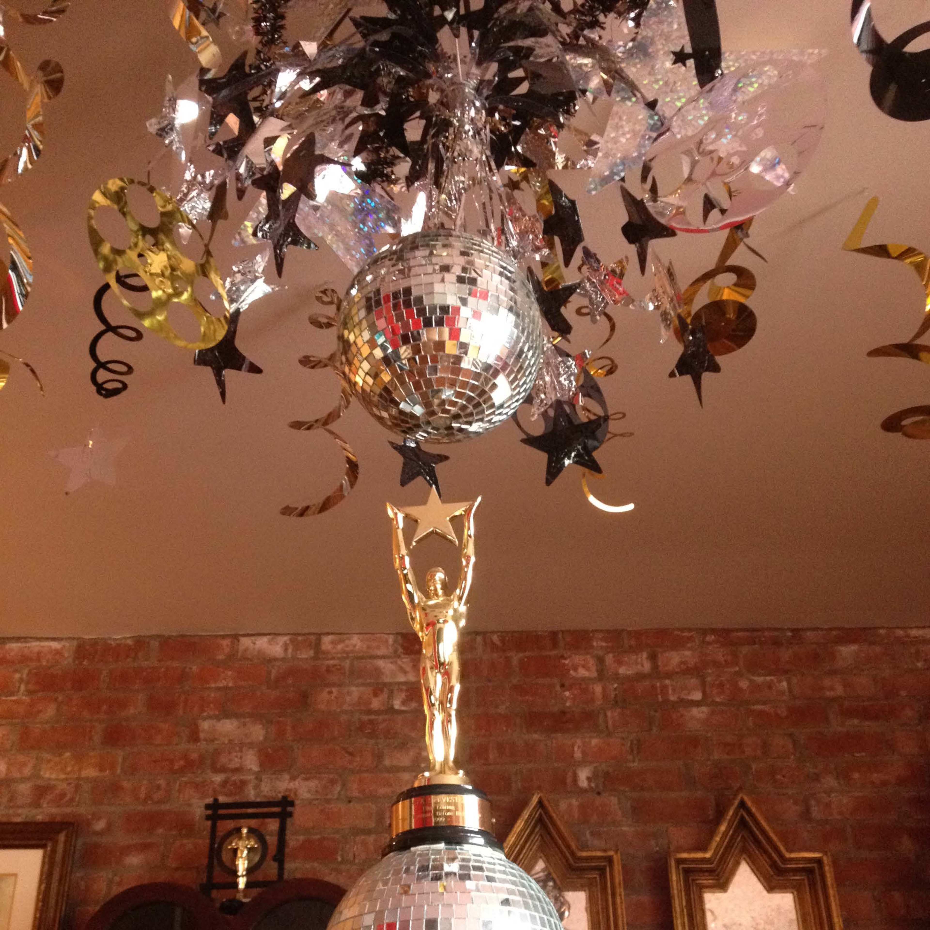 A party decoration featuring a gold trophy atop two mirrored disco balls, with colorful streamers hanging above.