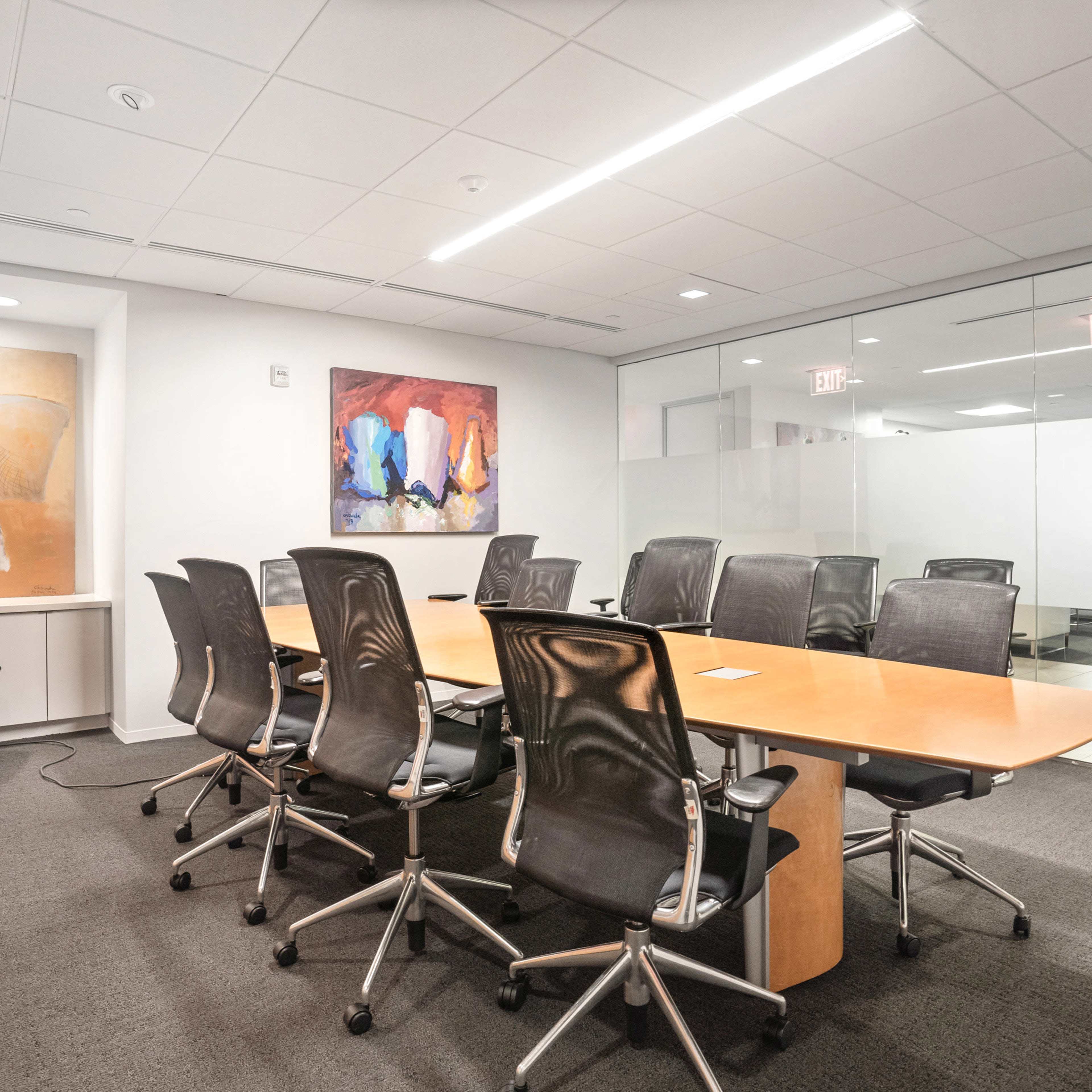 A modern conference room features a long wooden table surrounded by black office chairs, with two colorful abstract paintings on the walls.