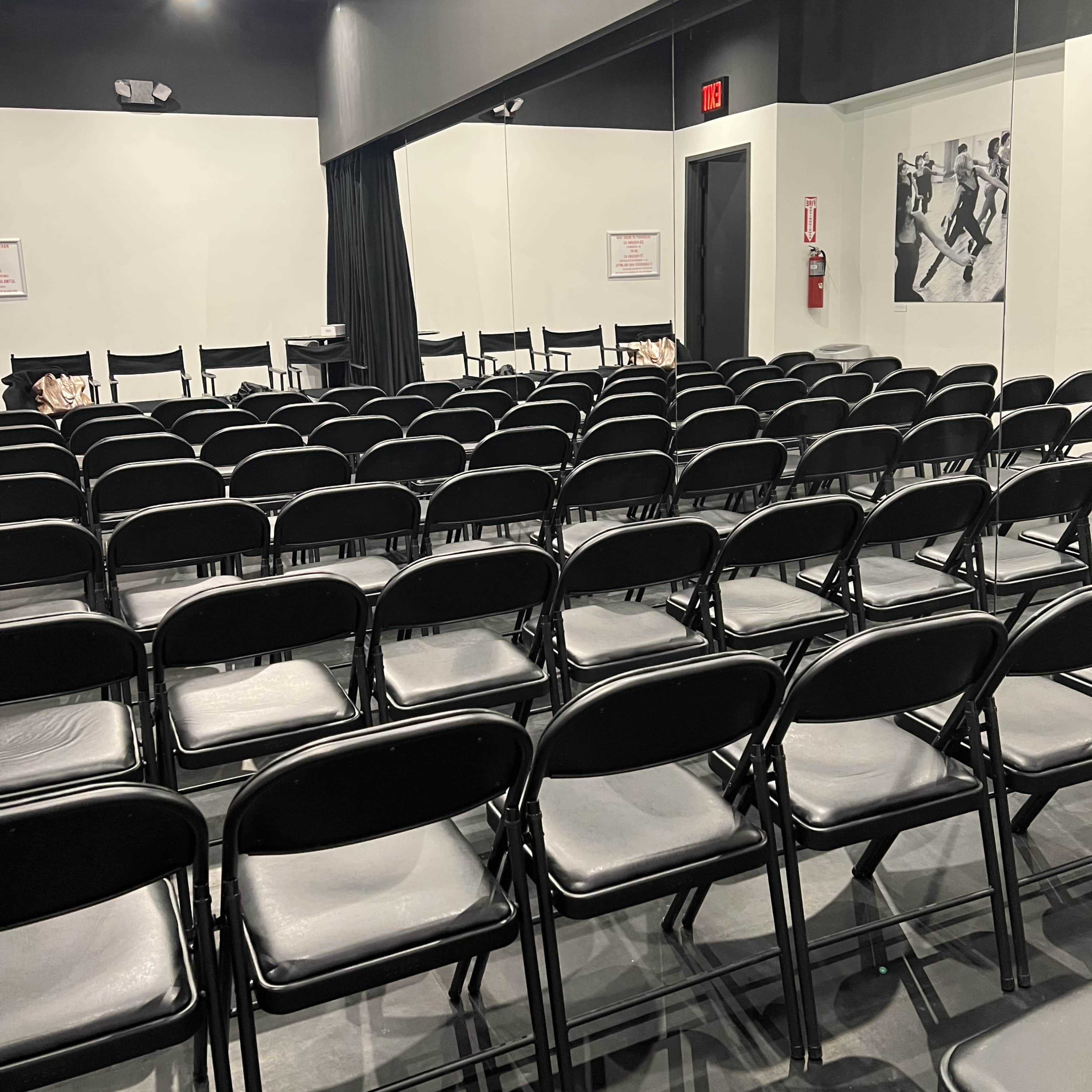 An empty room with rows of black folding chairs facing a wall, mirrored on one side.