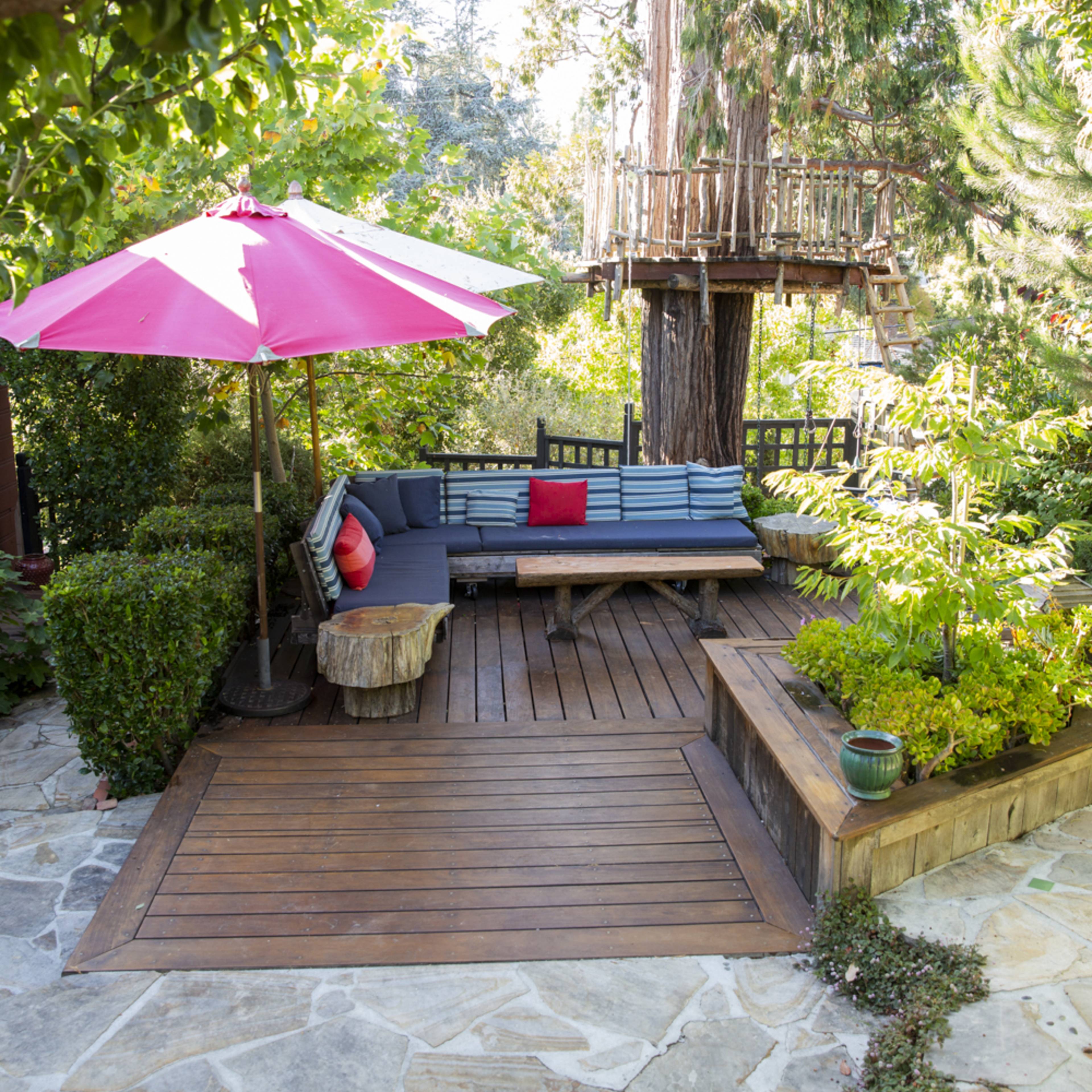A wooden patio with a seating area, a pink umbrella, and a tree house surrounded by greenery and stone pathways.