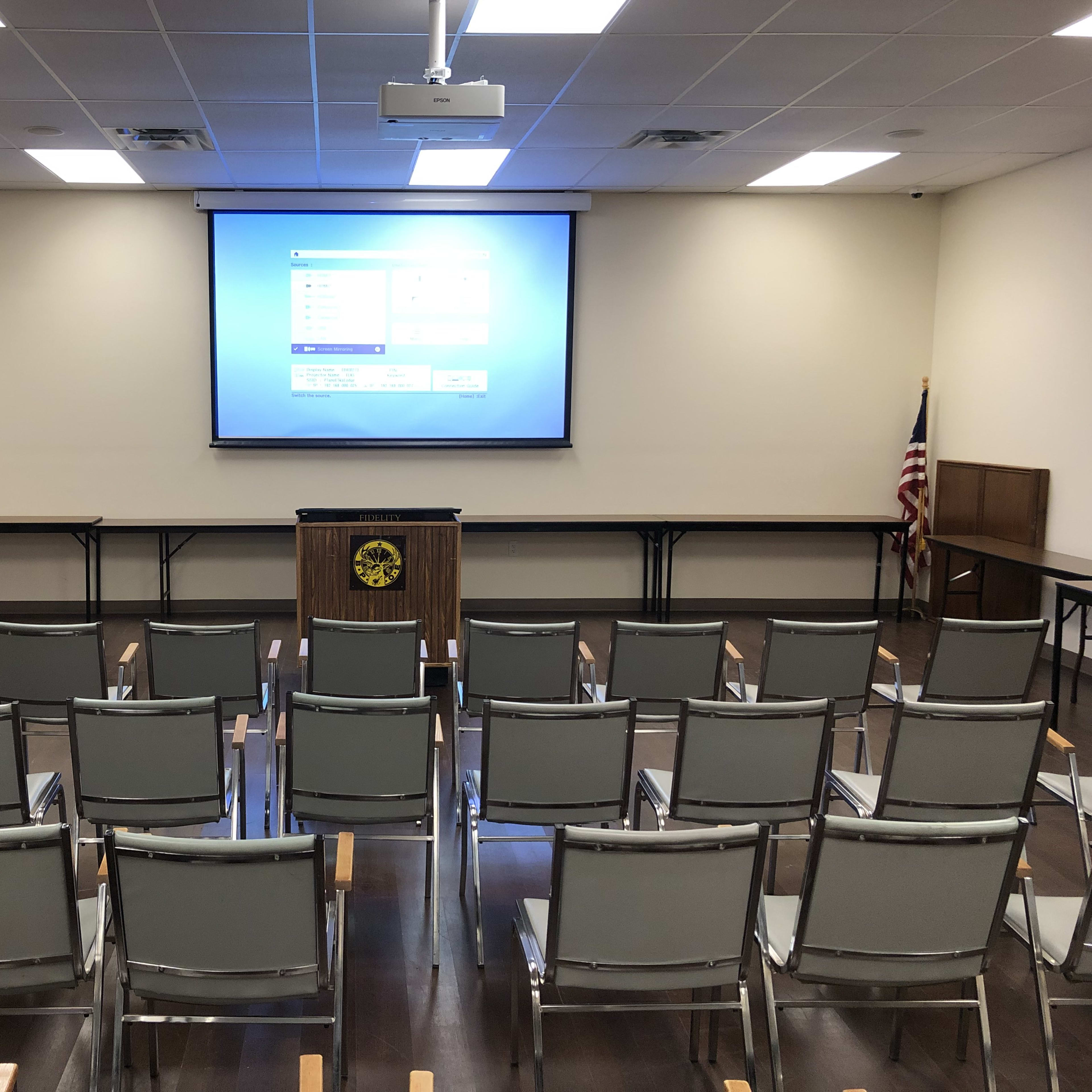 A meeting room is arranged with rows of gray chairs facing a large projection screen at the front.