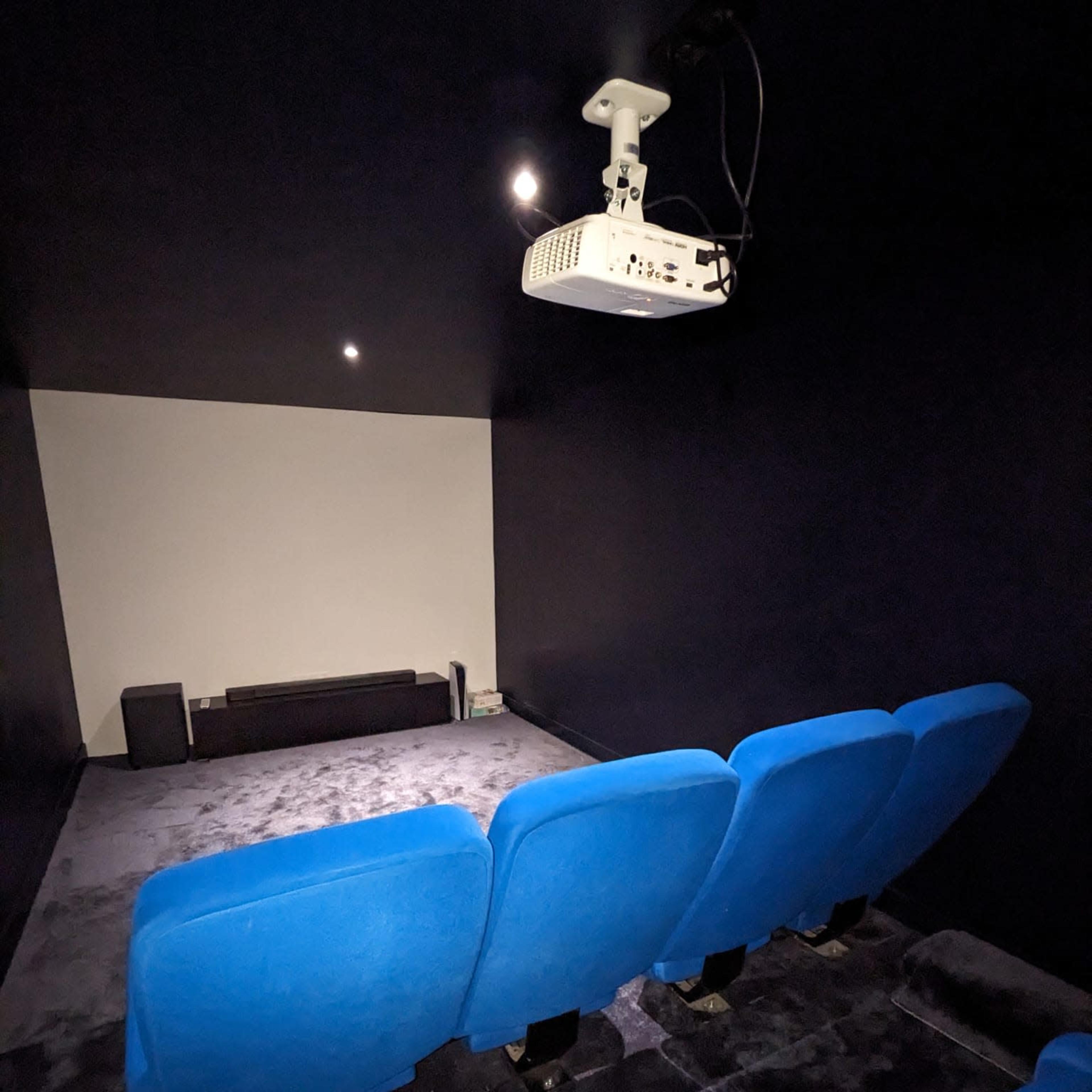 A small home theater room features blue seating, a projector mounted on the ceiling, and a blank screen against the wall.
