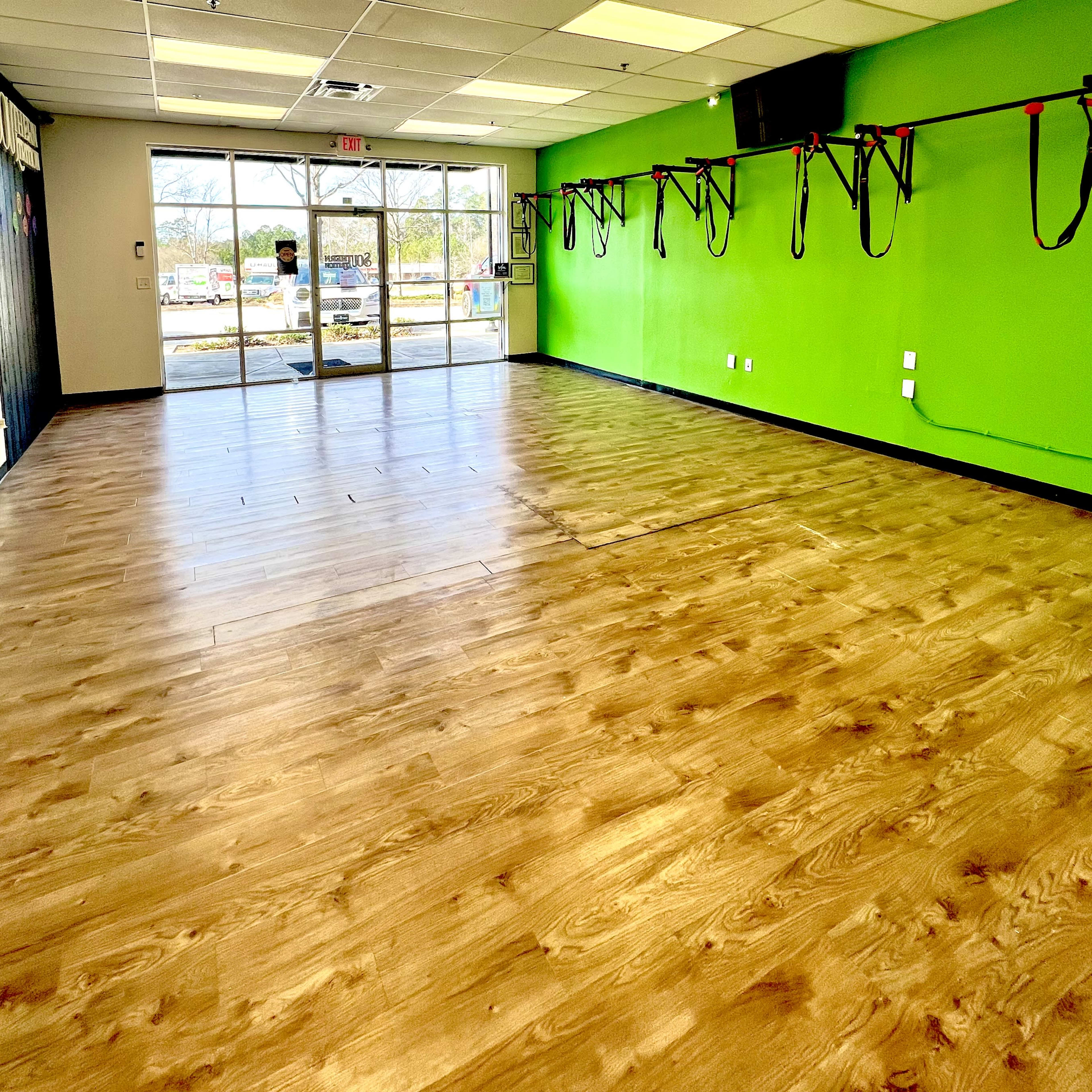 An empty fitness studio with wooden flooring, green walls, and suspension training equipment hanging on one side.