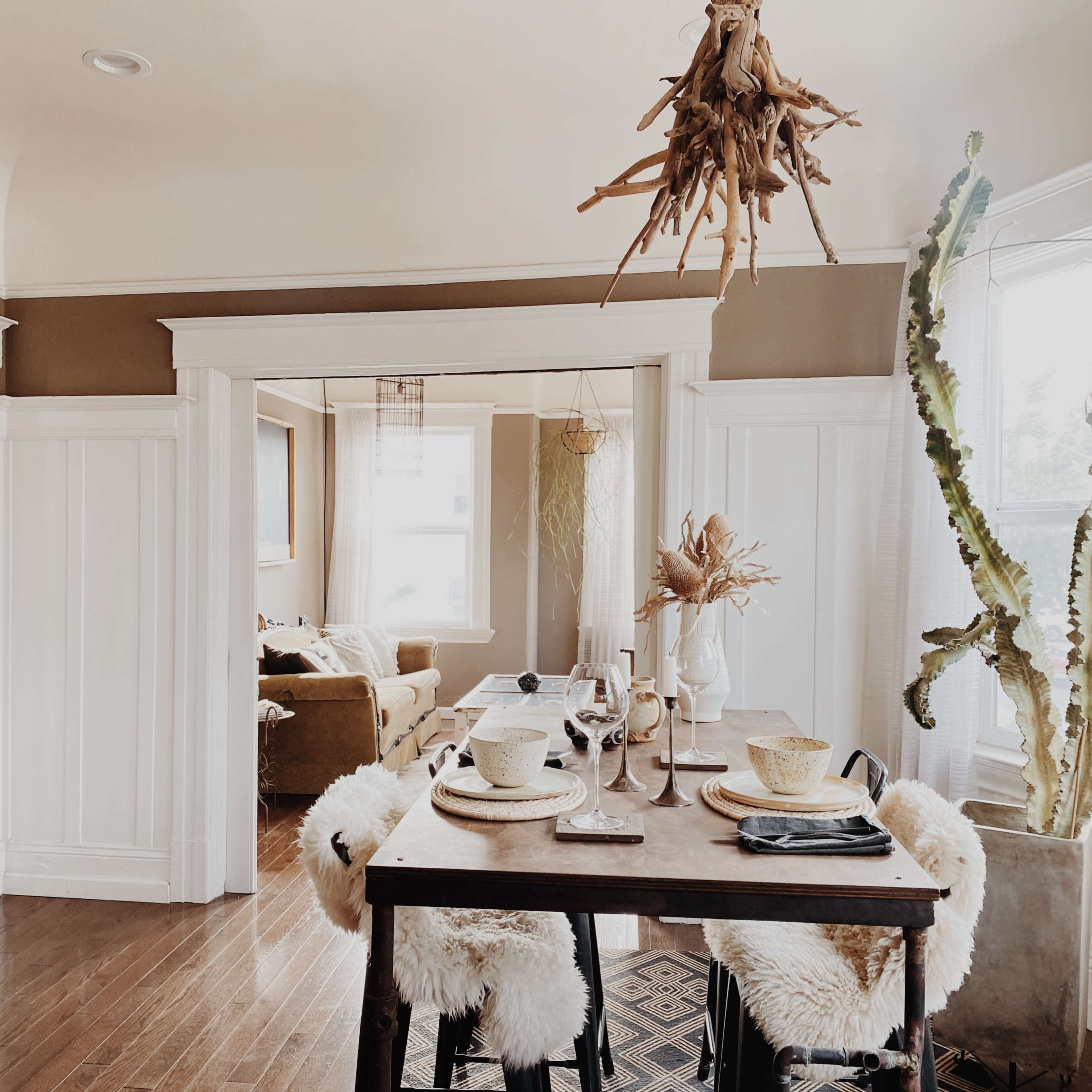 A dining area features a wooden table set with dishes, surrounded by chairs draped with fluffy covers, and leads into a cozy living room.