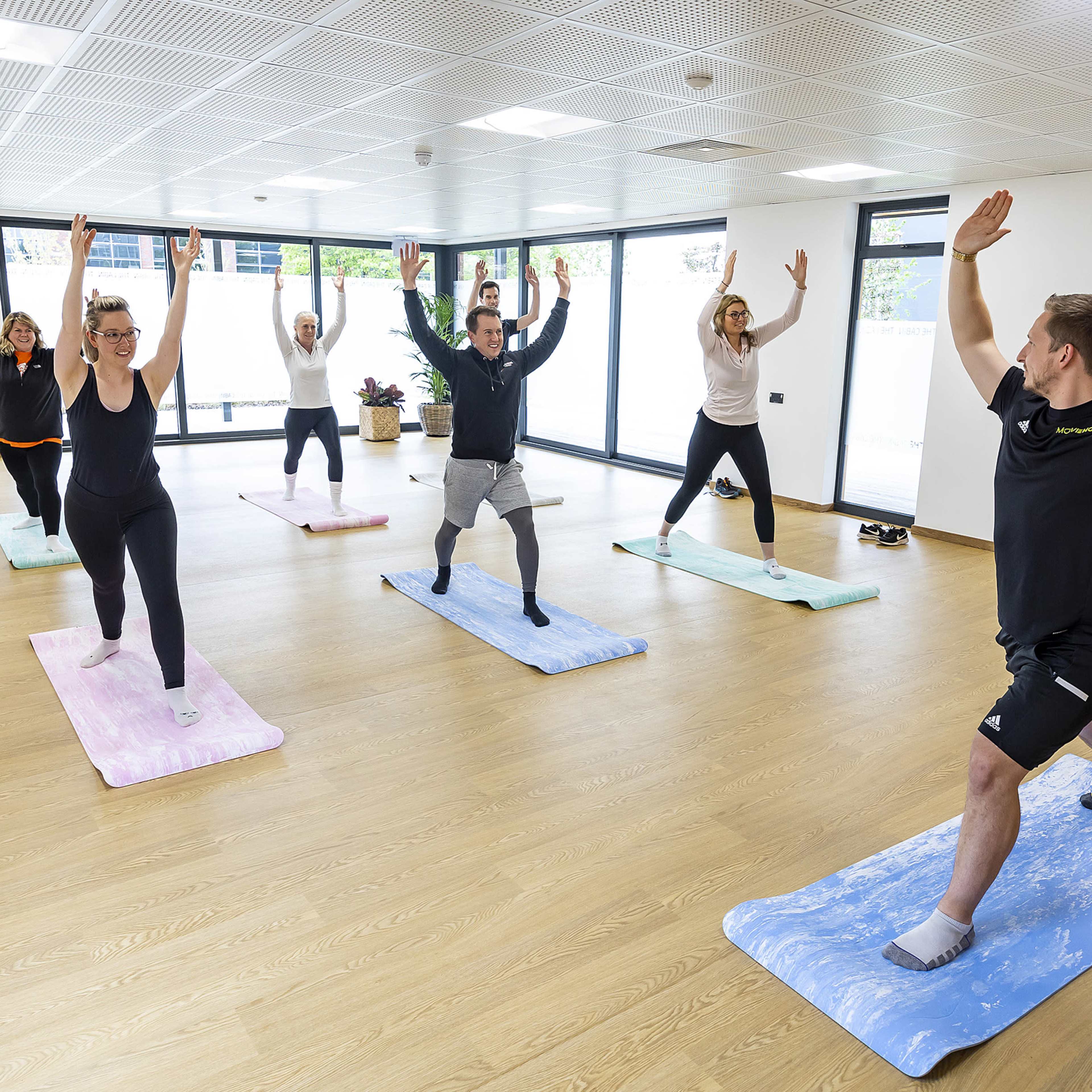 A group of six people is practicing yoga in a spacious studio, each using a mat while performing a pose with arms raised.