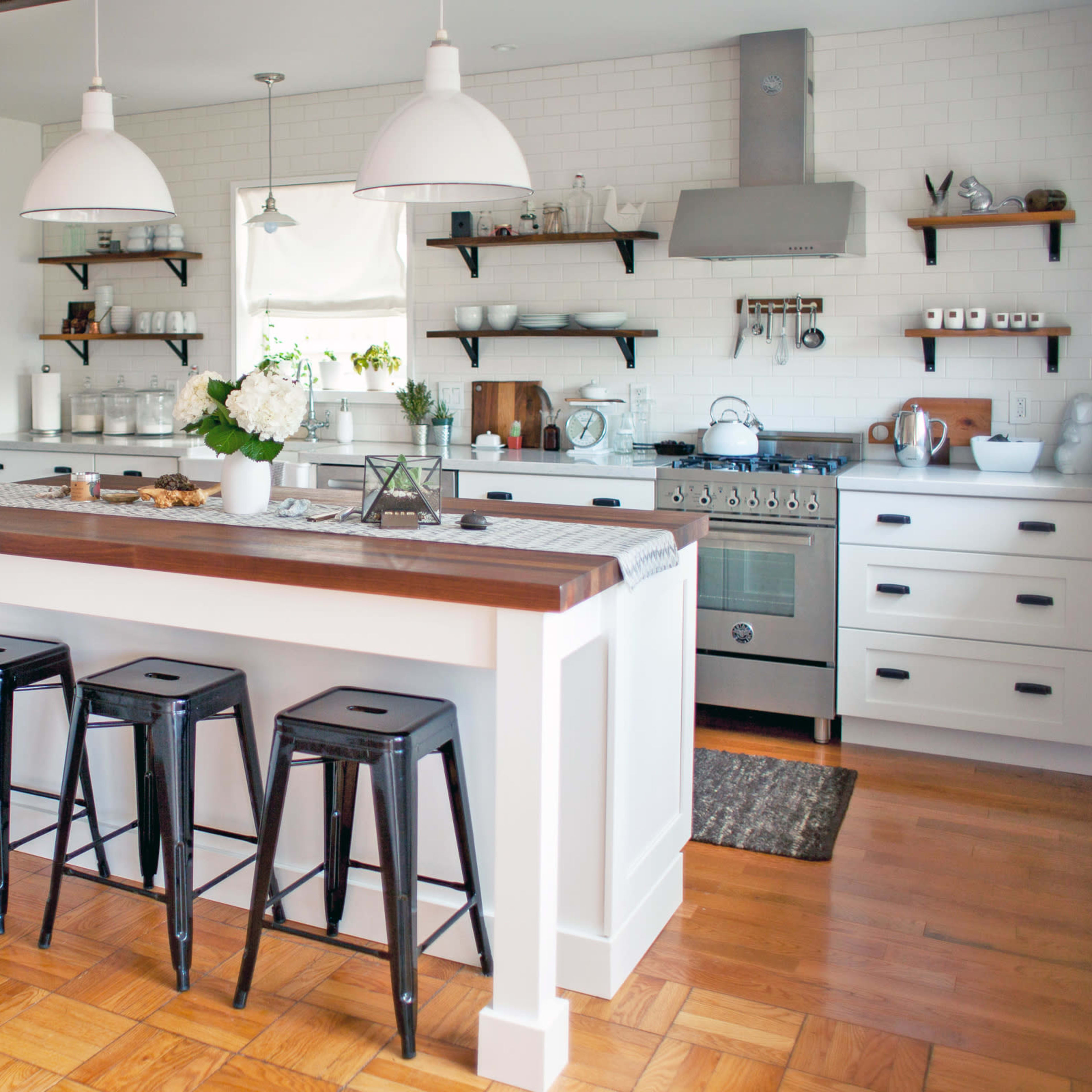 A modern kitchen with white cabinetry, a wooden island, and metal bar stools, featuring open shelves displaying dishware and a stove with a hood above.