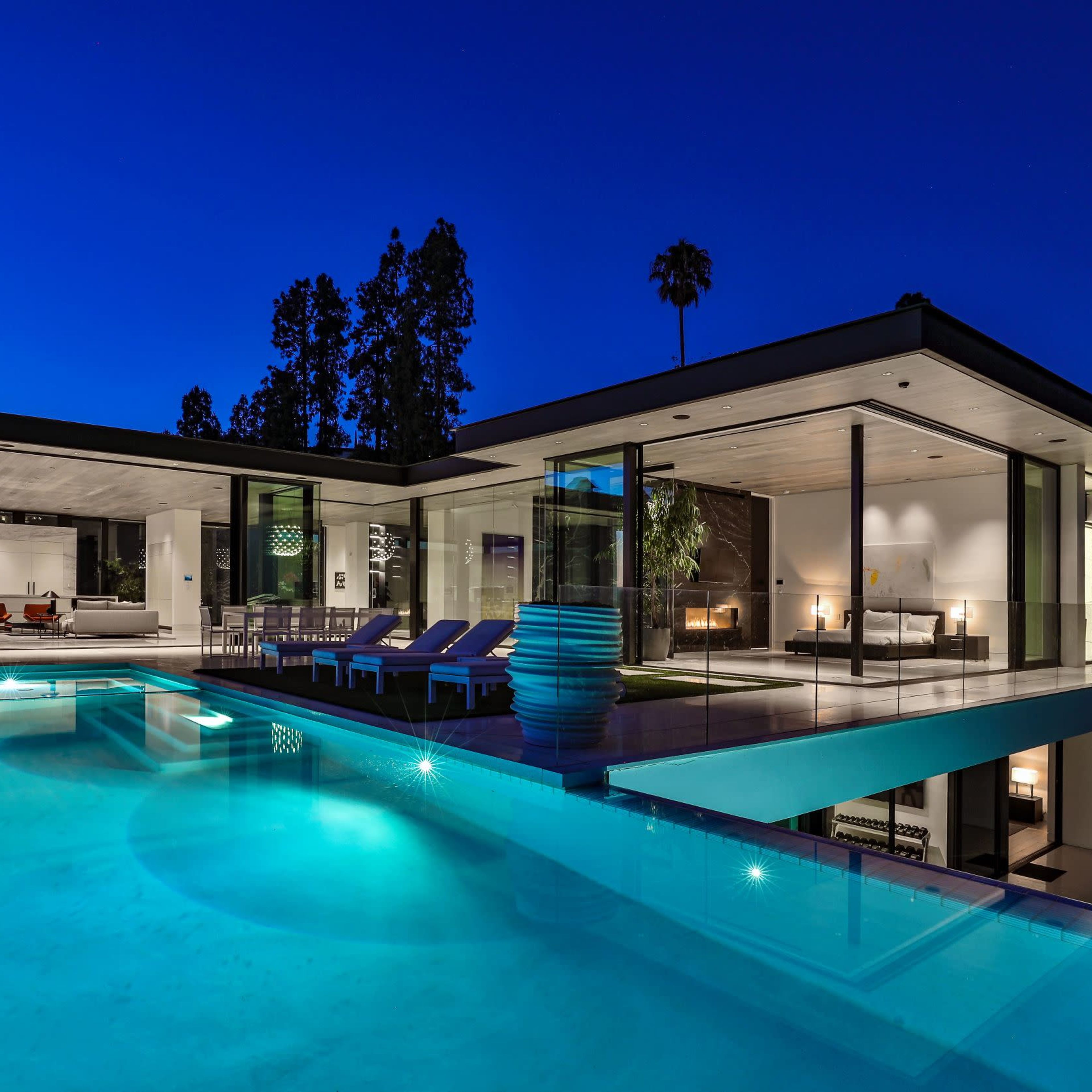 A modern home features a sleek pool area illuminated at night, with large glass walls revealing the luxurious interior.