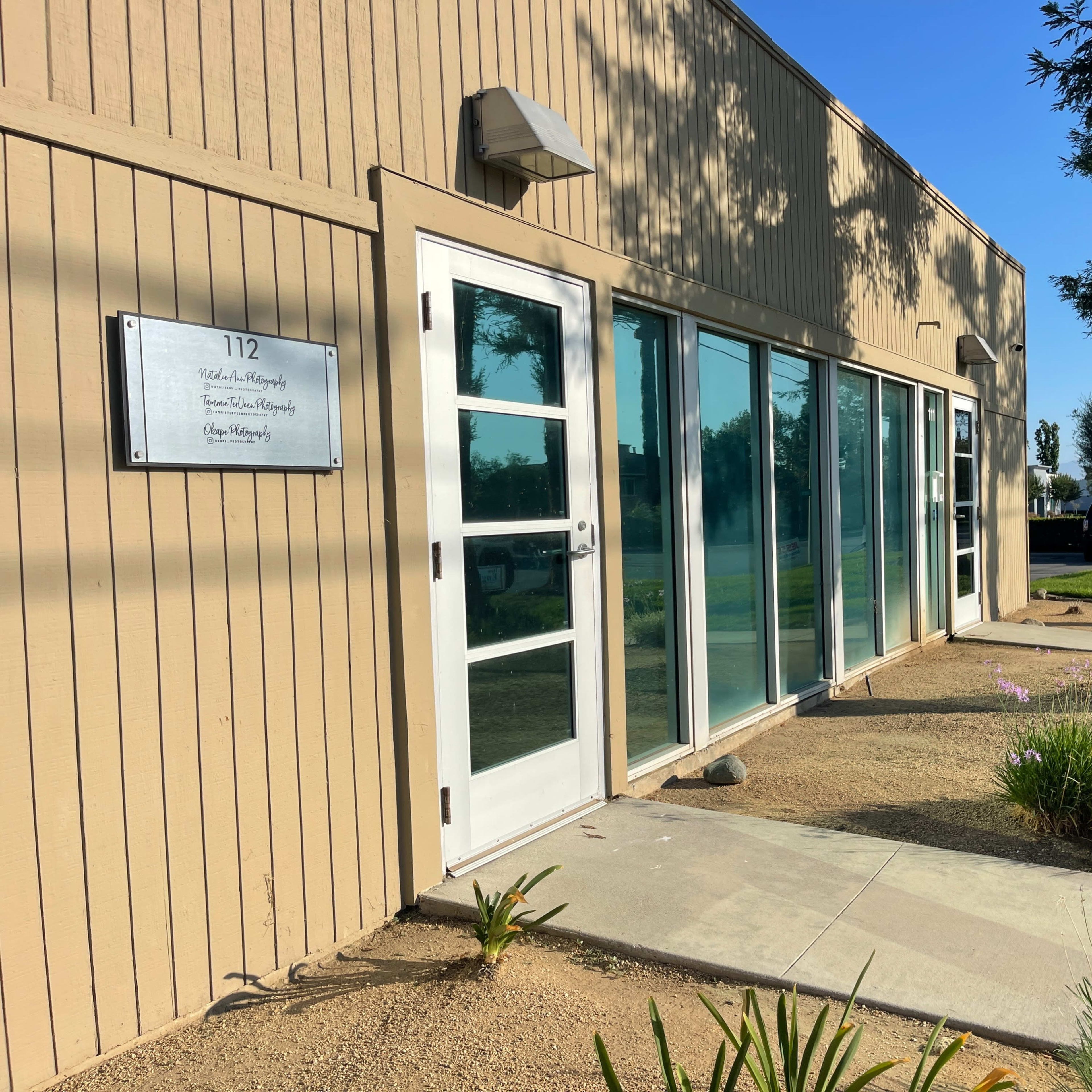 A light-colored building with large glass windows and a sign indicating the address, surrounded by a landscaped area with plants.