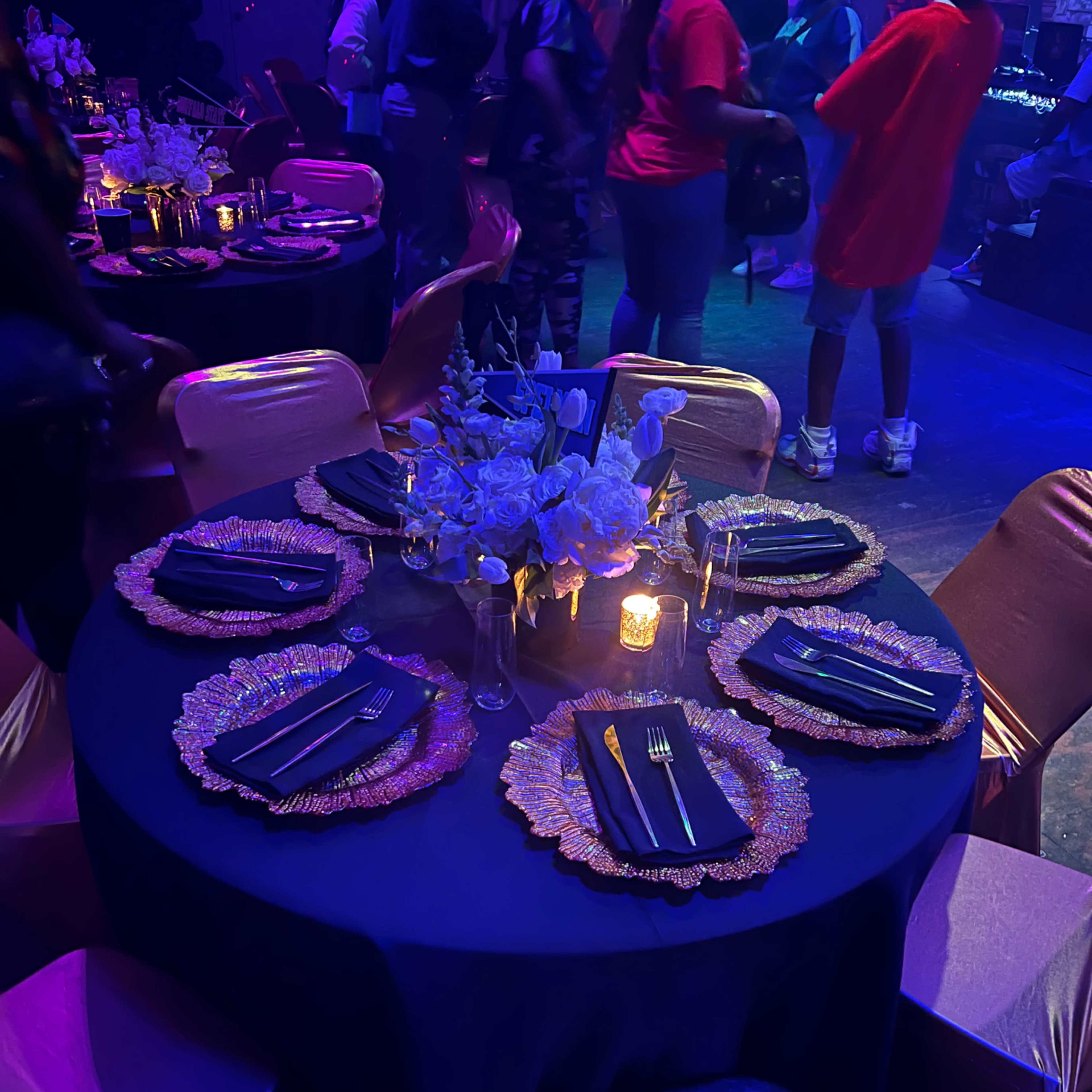 A round table is elegantly set with dark plates and sparkling chargers, surrounded by gold and pink chairs, and decorated with flowers and a candle.