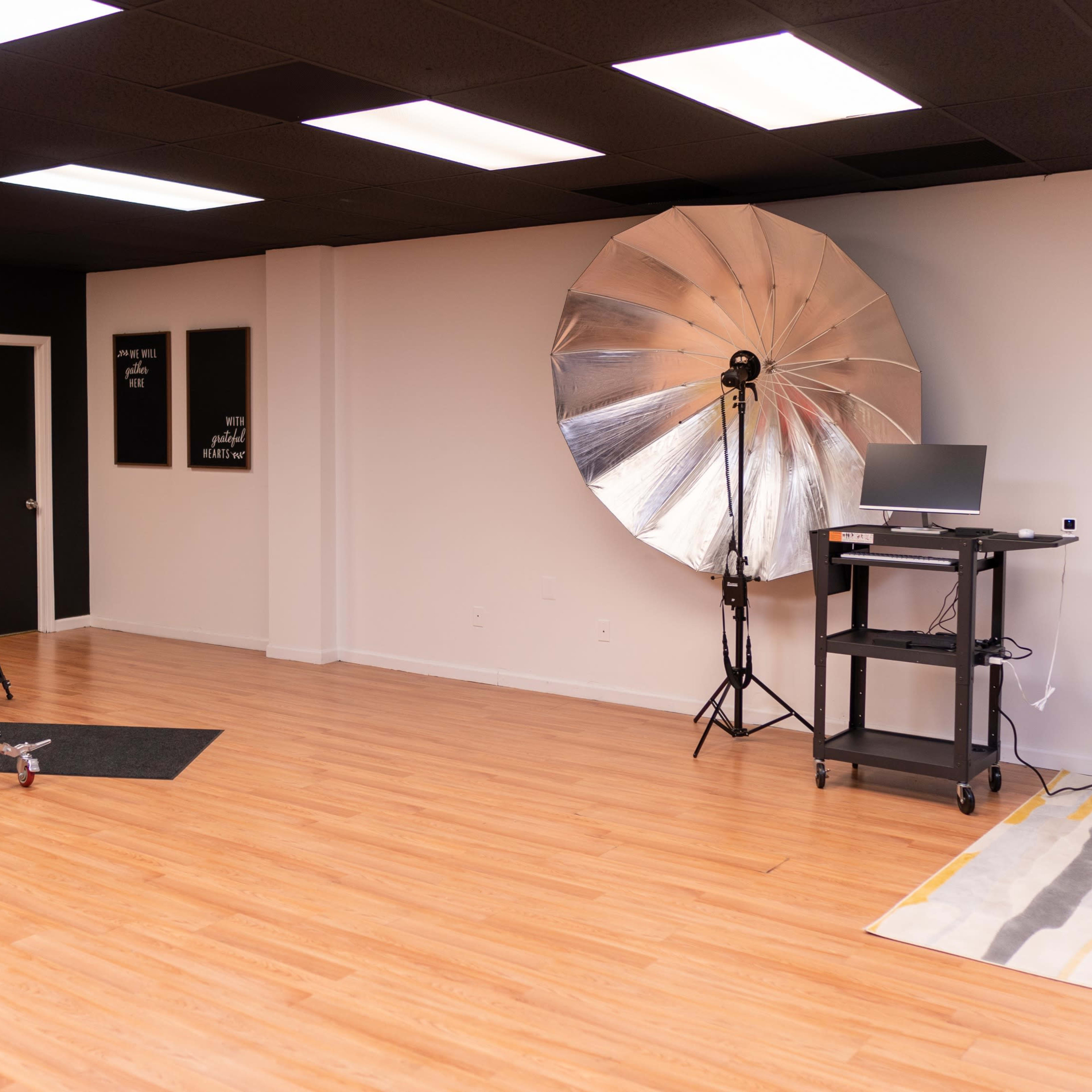 A spacious photography studio featuring a large silver reflector, a softbox light, a computer station, and various props arranged on a wooden floor.