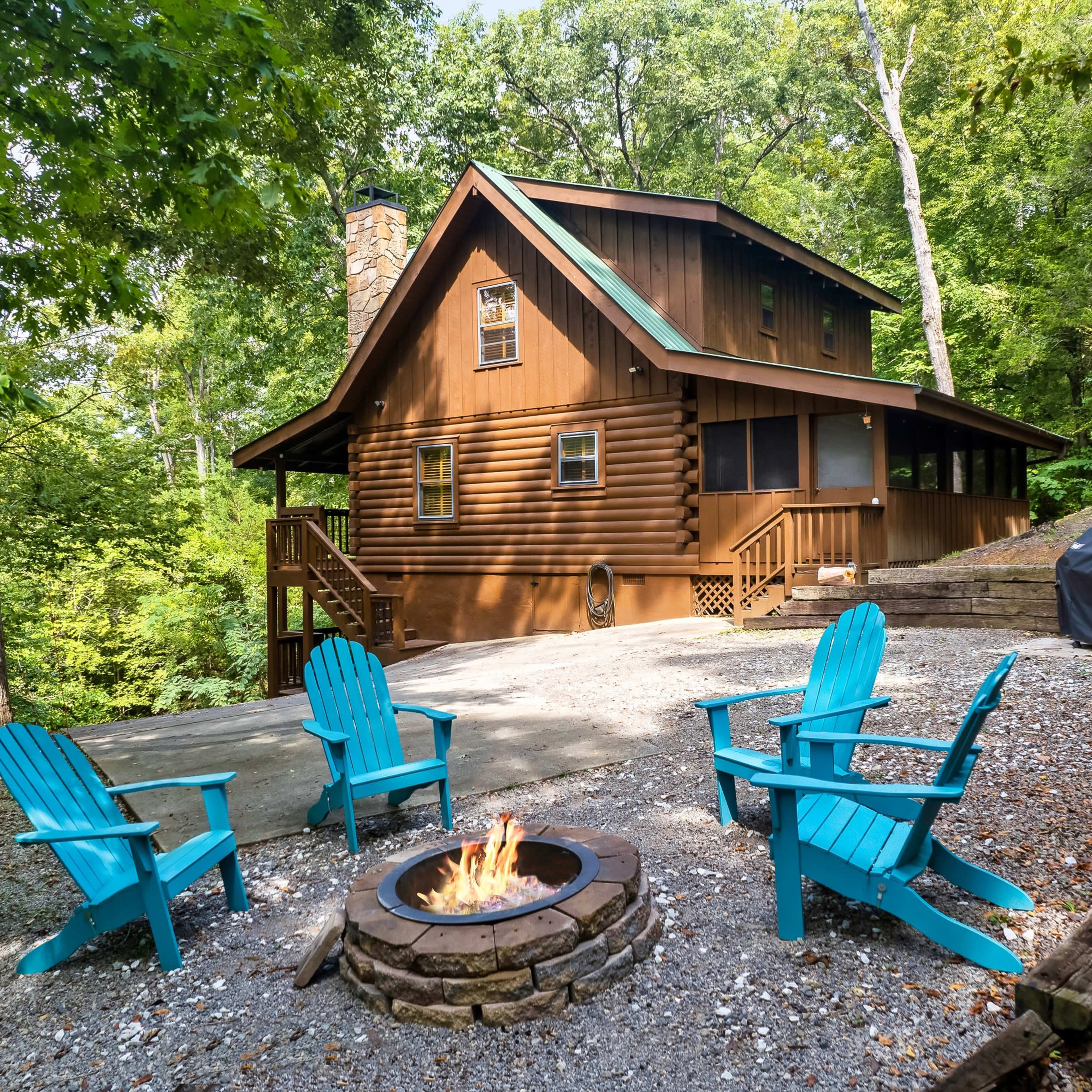 A two-story log cabin sits nestled among trees, with a fire pit and blue Adirondack chairs arranged around it in a gravel area.