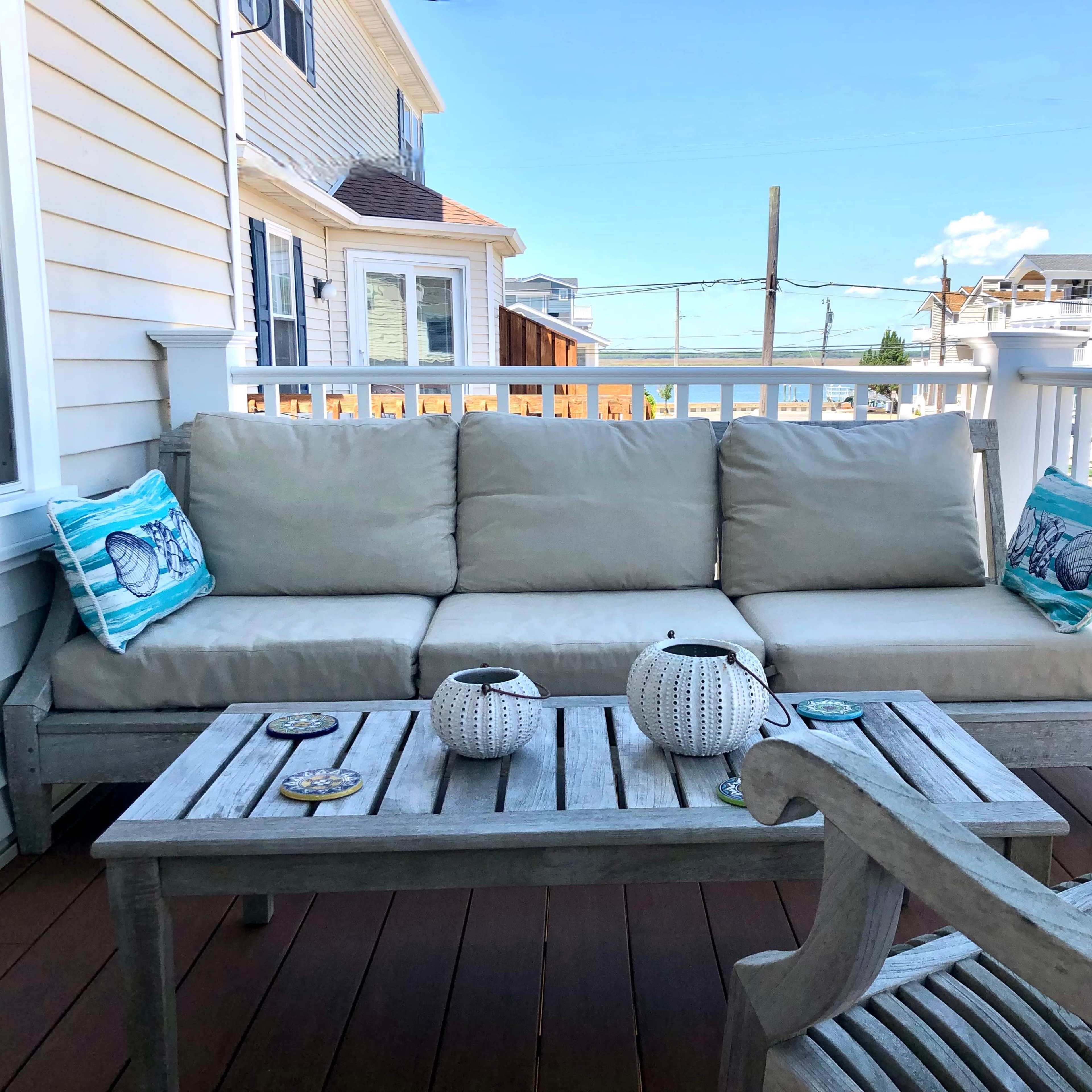 A cozy outdoor seating arrangement features a beige couch with decorative pillows, a wooden table with two lanterns, and a clear blue sky in the background.