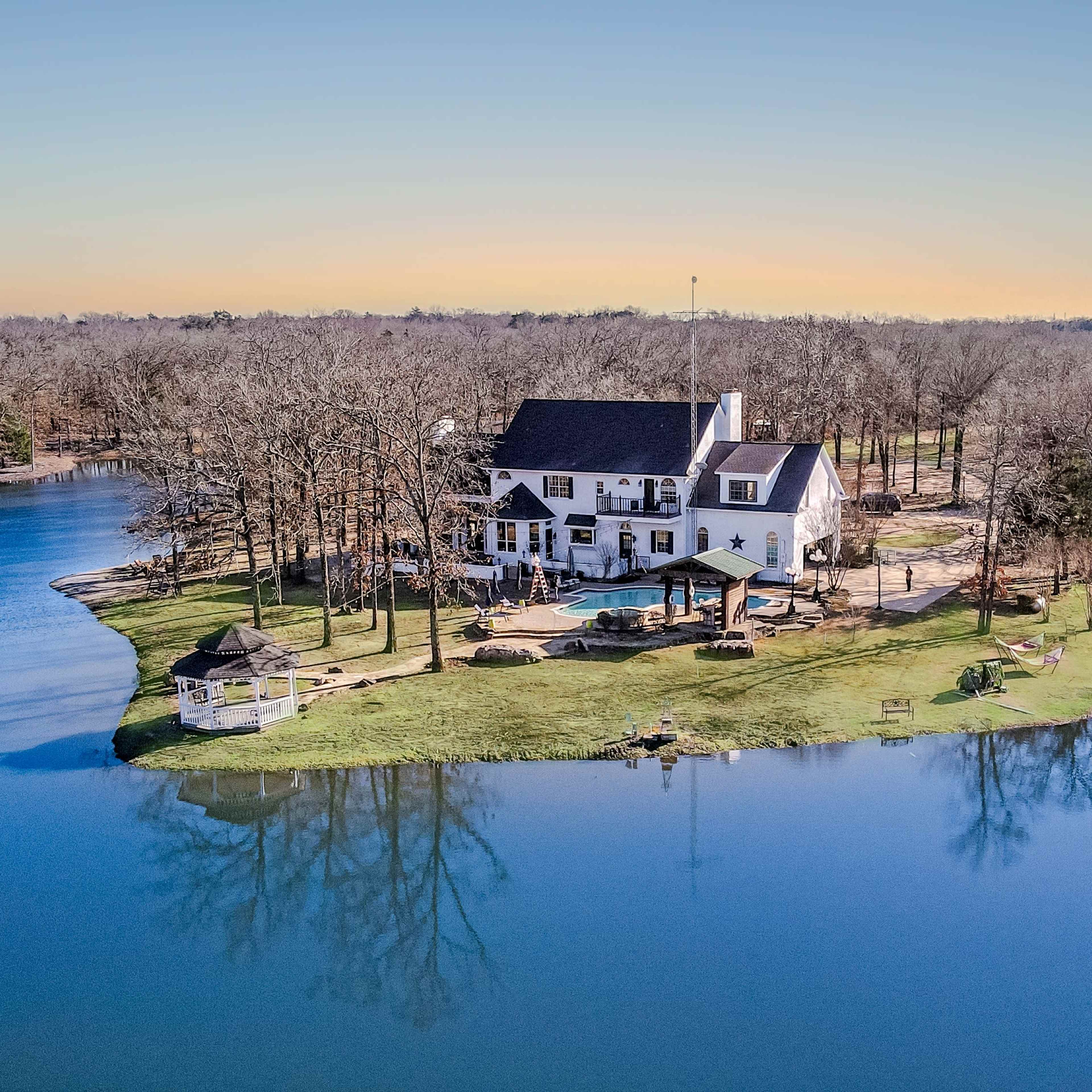 A house is situated on a small island surrounded by calm water and trees in the background.