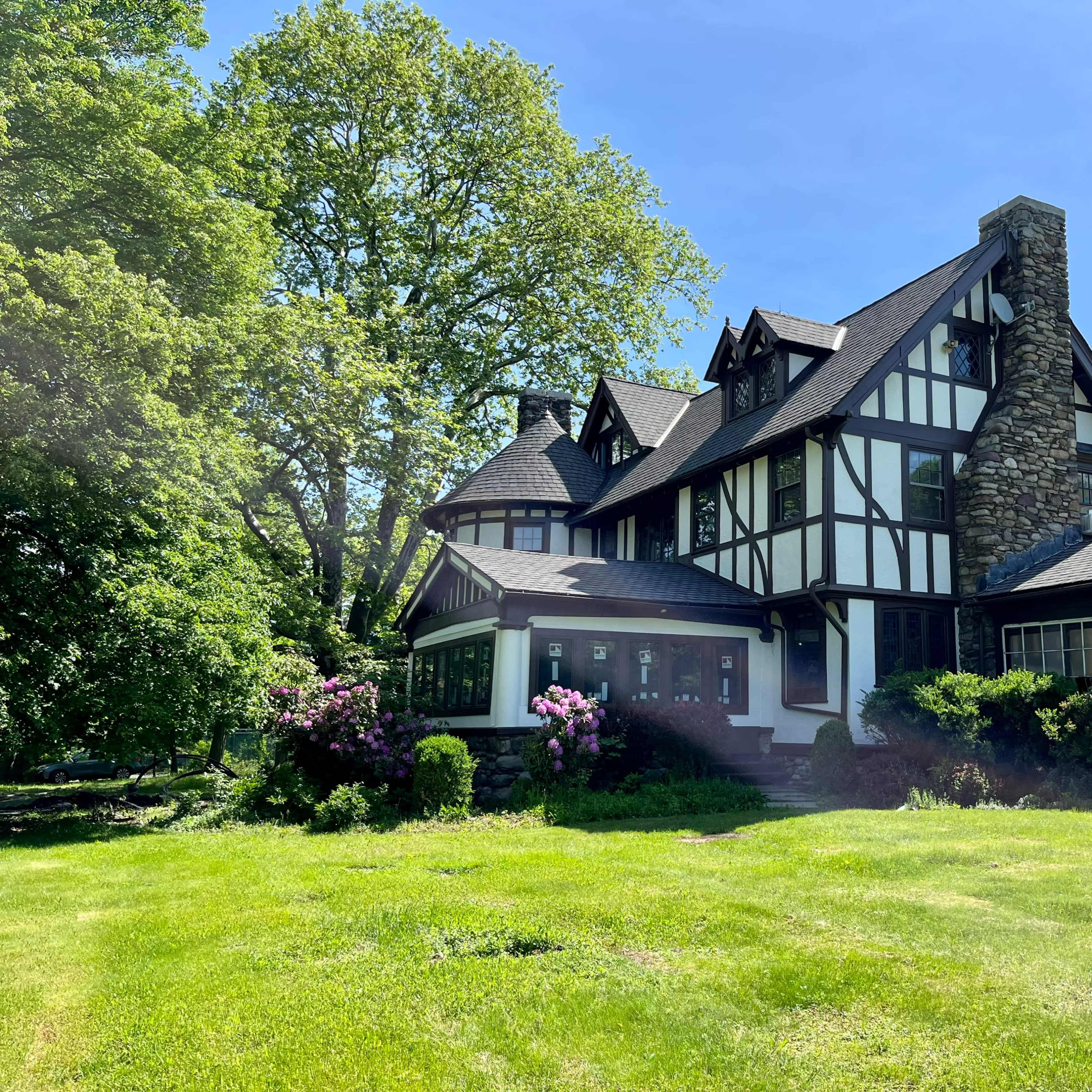 A large, two-story Tudor-style house with stone and timber accents is surrounded by lush green grass and trees.