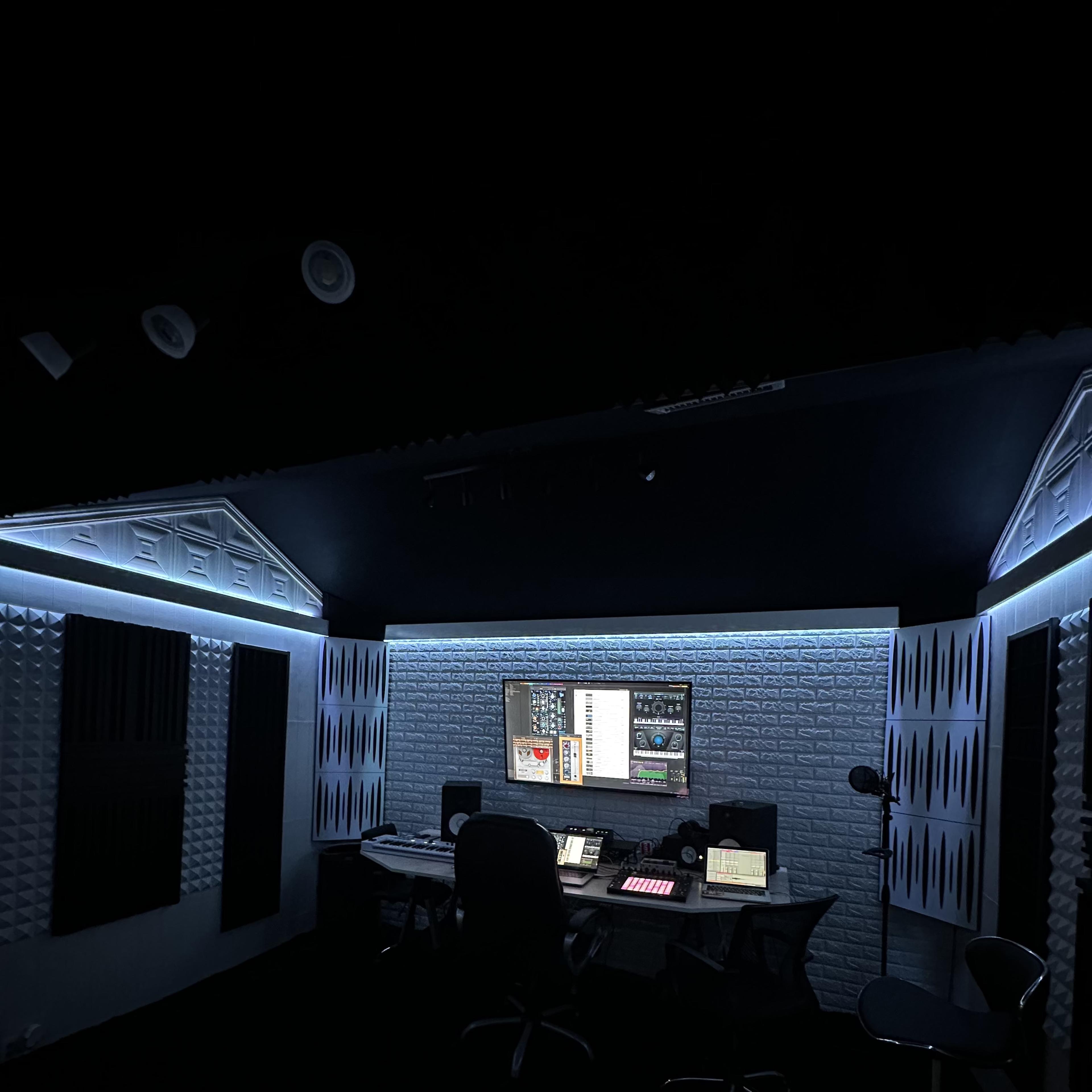 A dimly lit recording studio features a large desk with audio equipment, surrounded by soundproof wall panels.