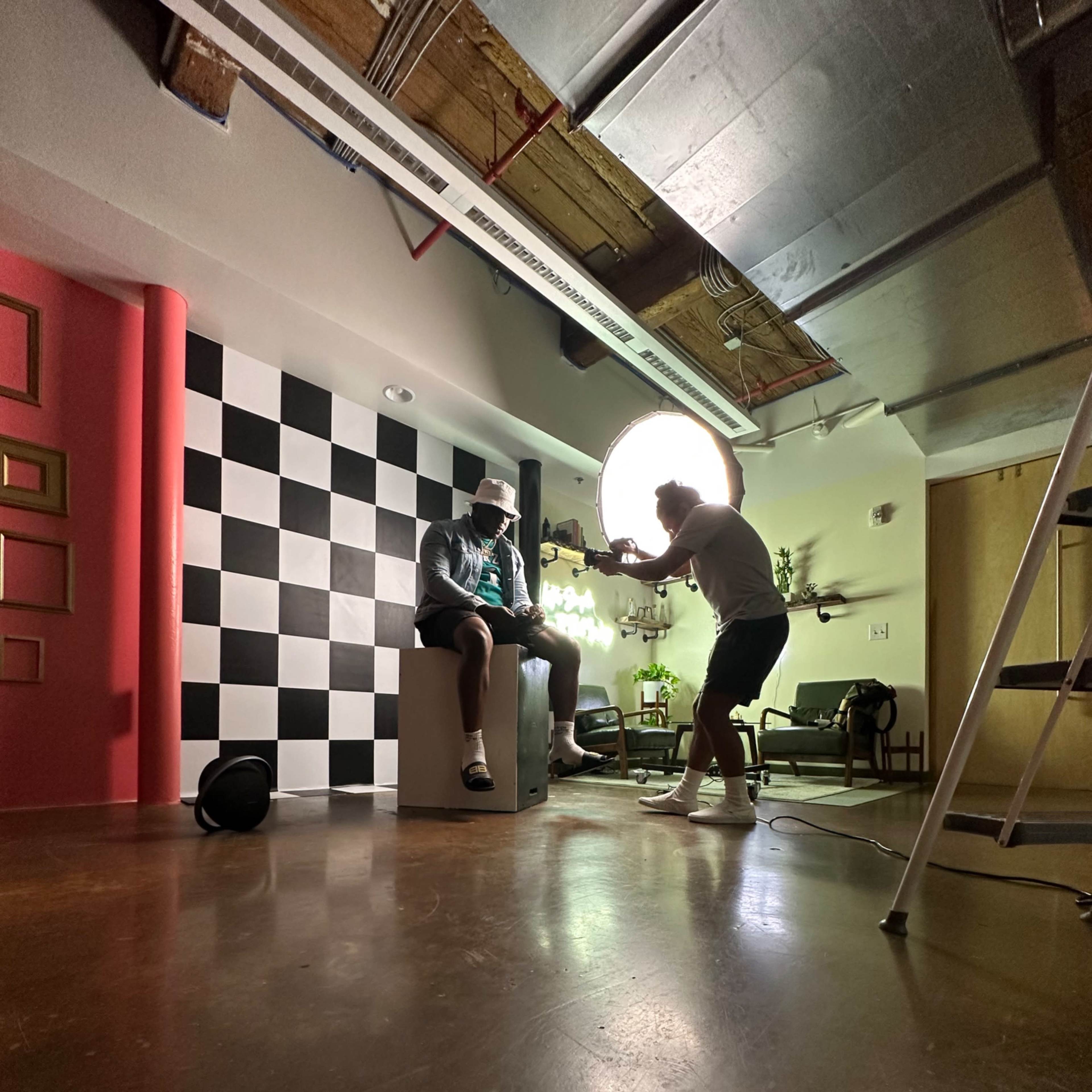 A photographer is capturing an indoor photo shoot featuring a model seated on a pedestal in a studio with a checkered backdrop and a large light source.