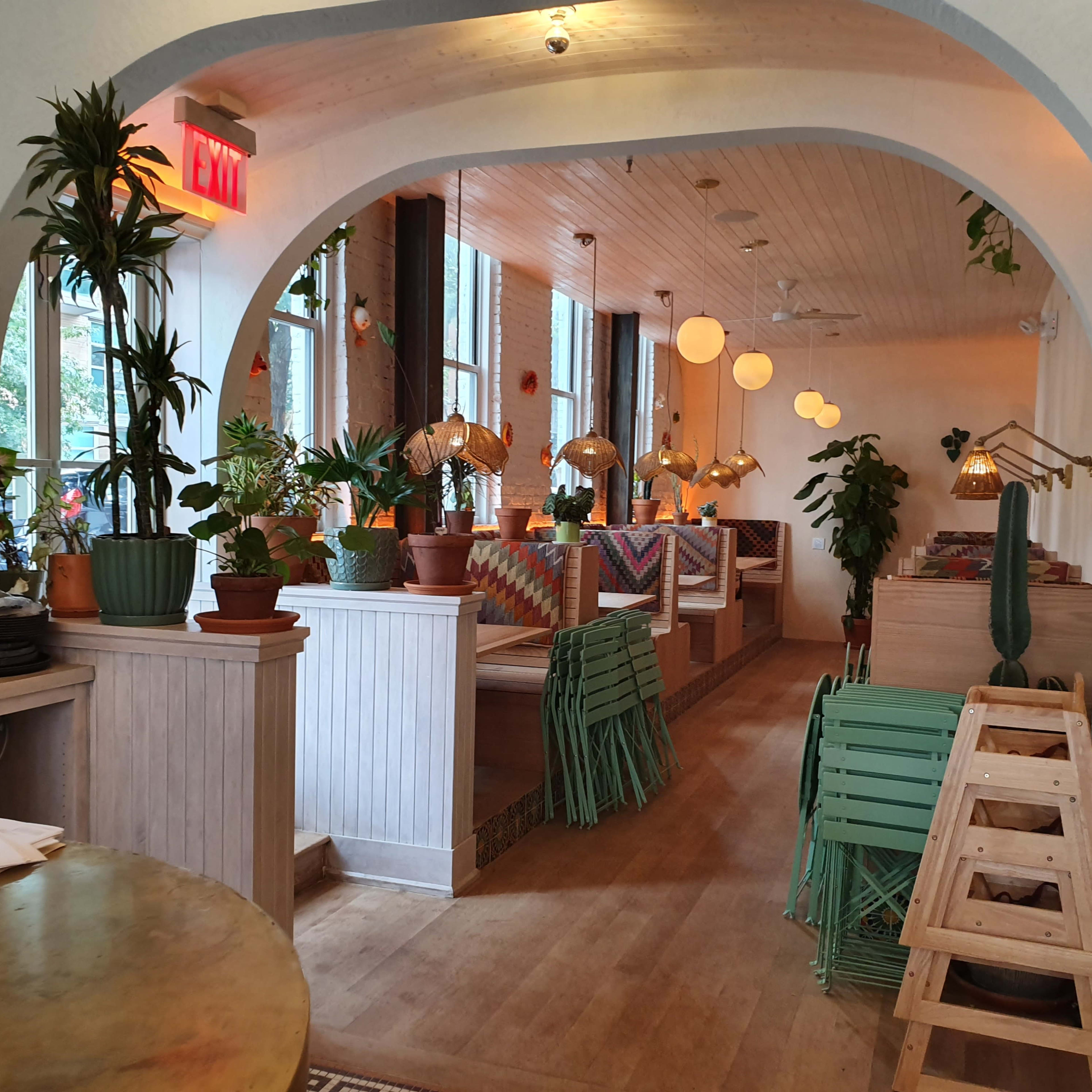 A brightly lit café interior featuring arched doorways, wooden ceiling, potted plants, and a mix of seating arrangements with decorative lighting.