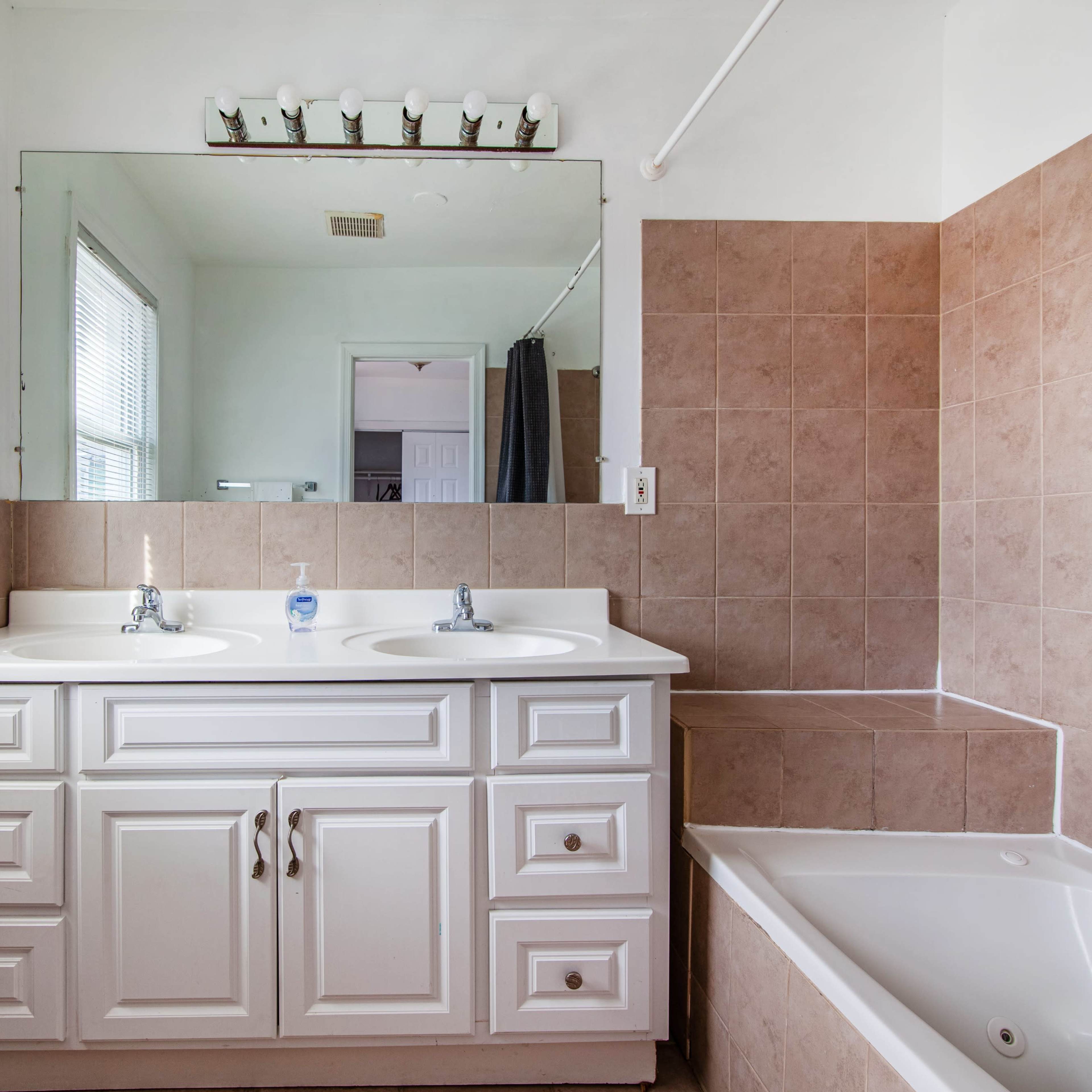 A bathroom with a double vanity, large mirror, and a bathtub, featuring beige tiled walls and white cabinetry.
