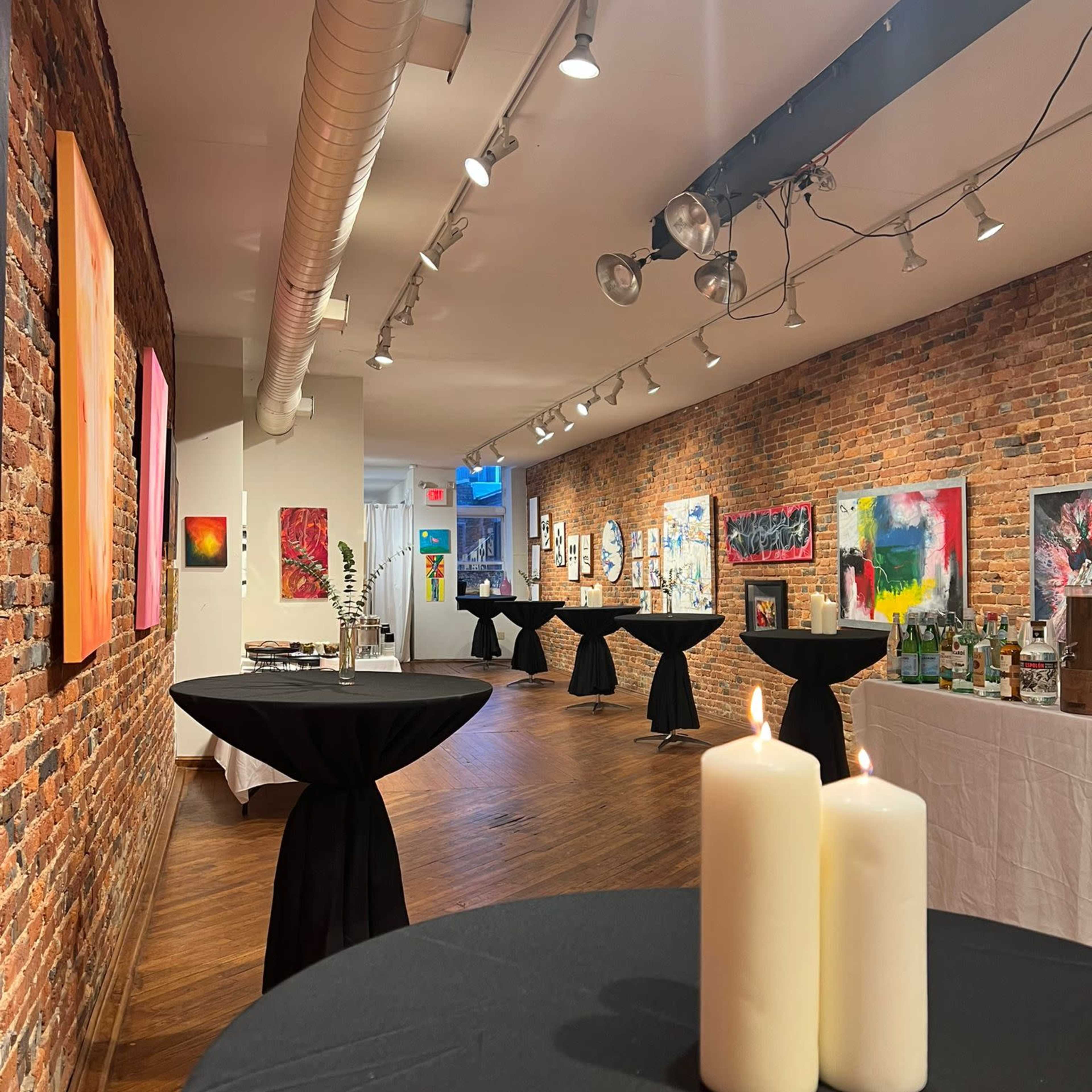 An art gallery featuring brick walls, black tables with candles, and various artworks displayed on the walls.