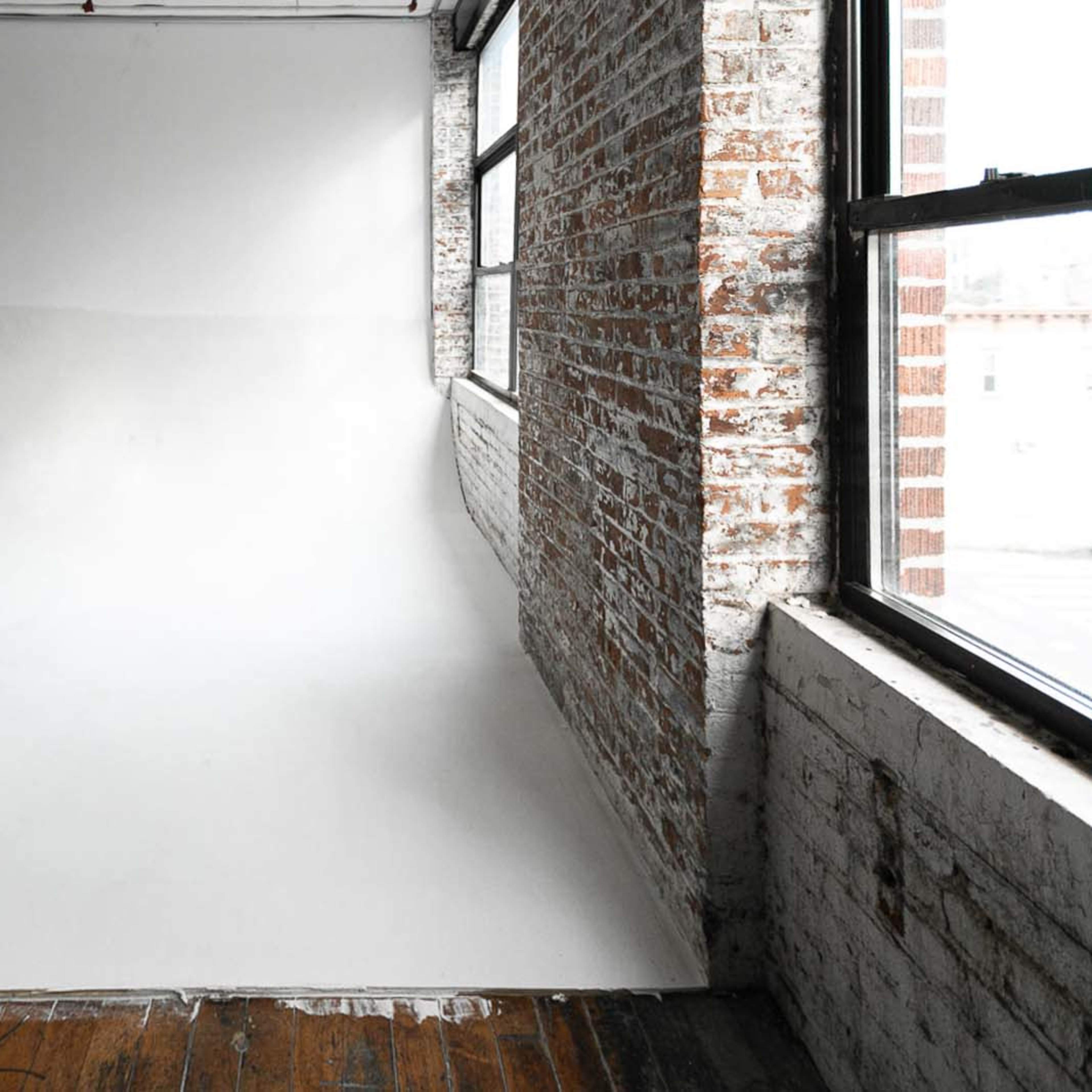 A spacious studio with a curved white backdrop and exposed brick walls, illuminated by natural light from large windows.