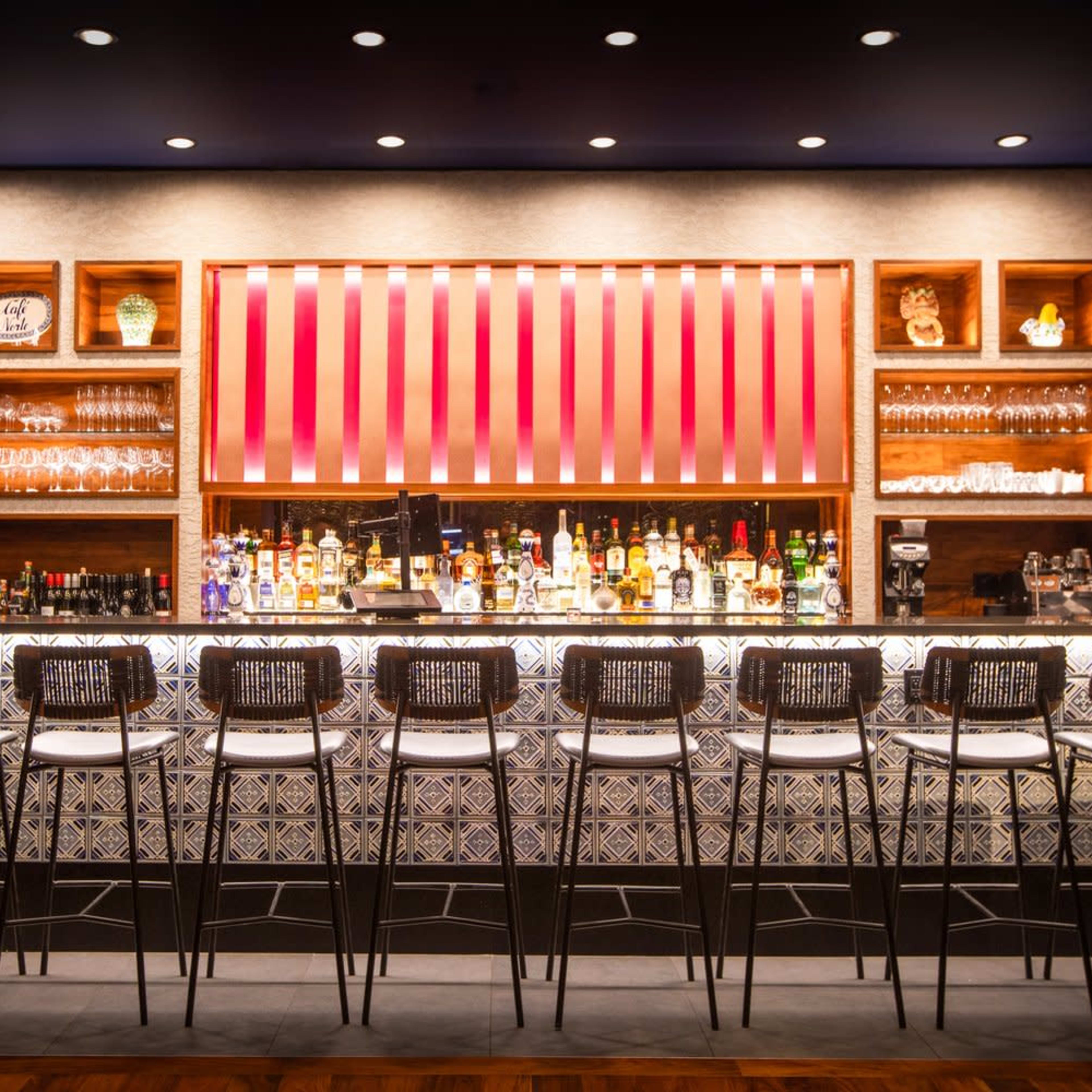 A modern bar features a long counter with high-backed stools in front of a well-stocked display of colorful liquor bottles and glassware against a stylish backdrop.