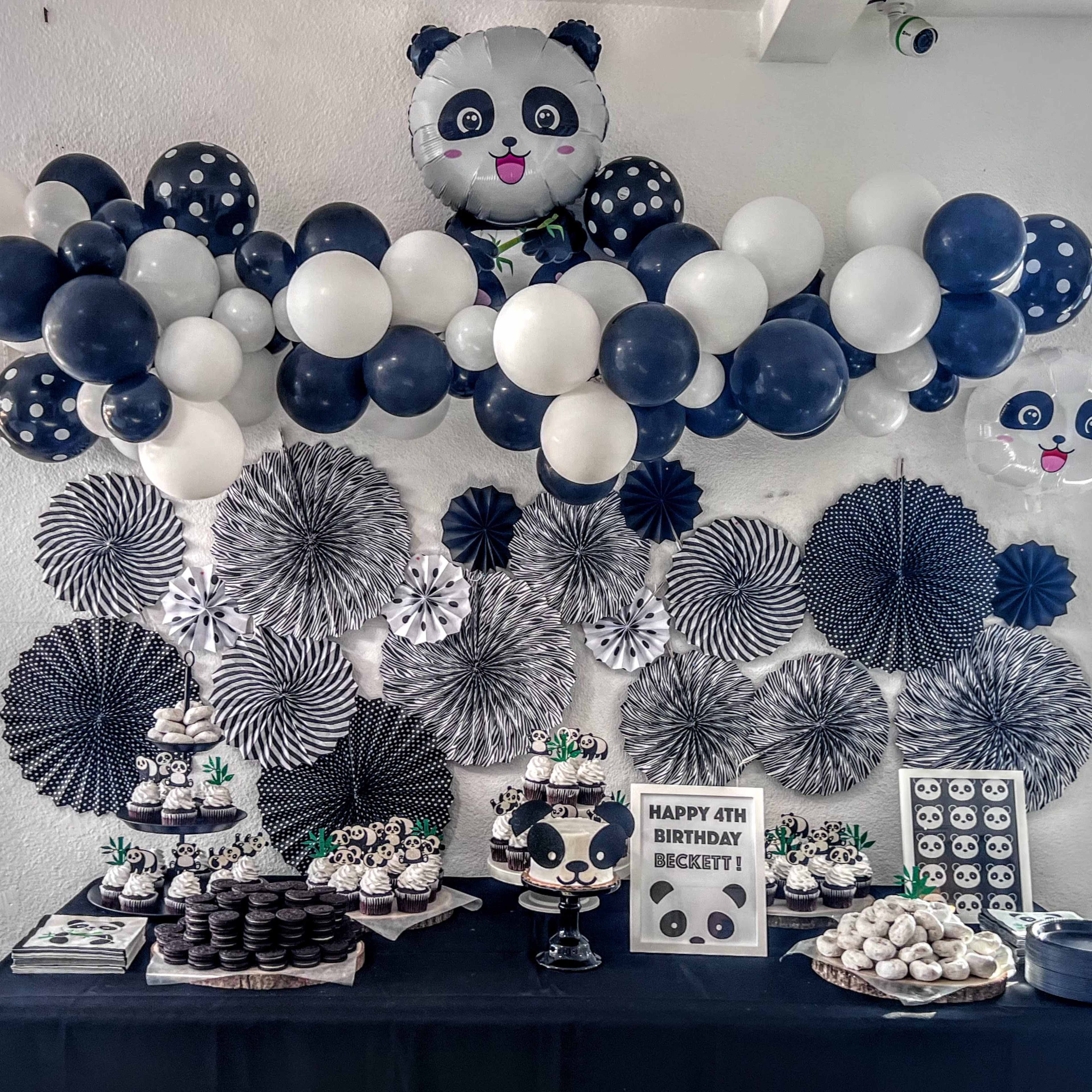 A festive birthday party setup features a table adorned with panda-themed decorations, a cake, and assorted treats, all complemented by a backdrop of balloons and paper fans.