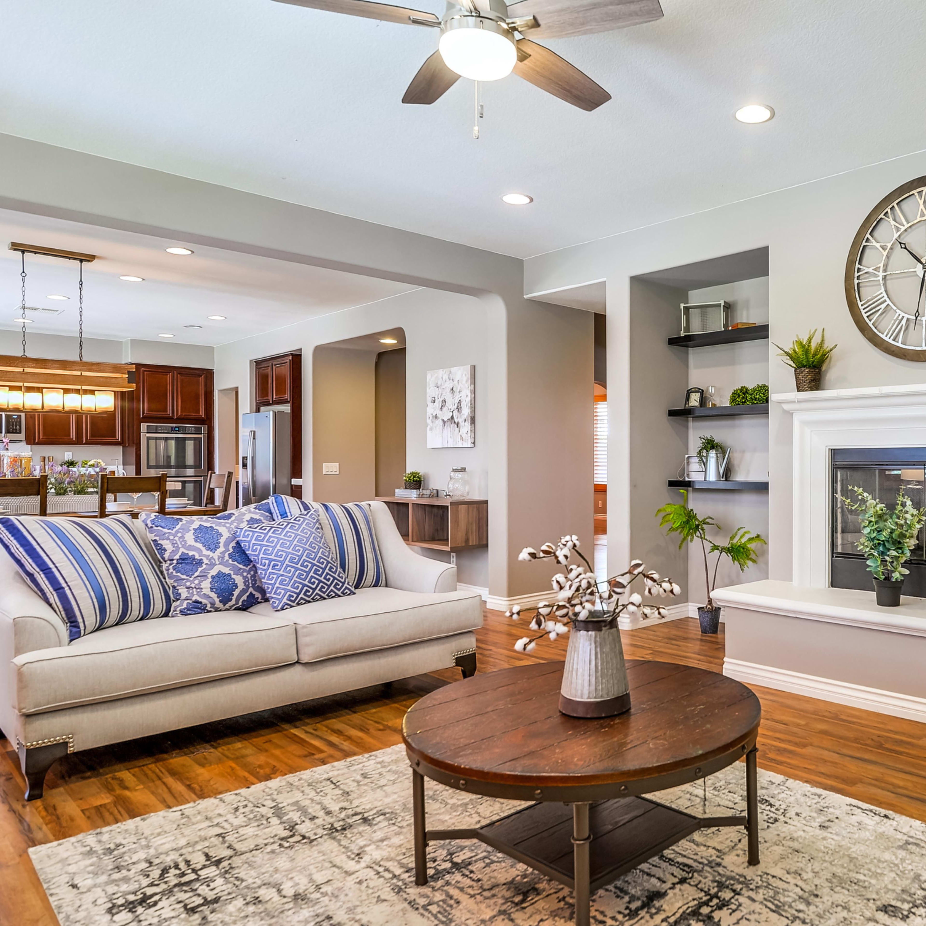 A living room features a white fireplace, a grey wall clock, a round wooden coffee table, and a beige sofa with blue striped throw pillows.