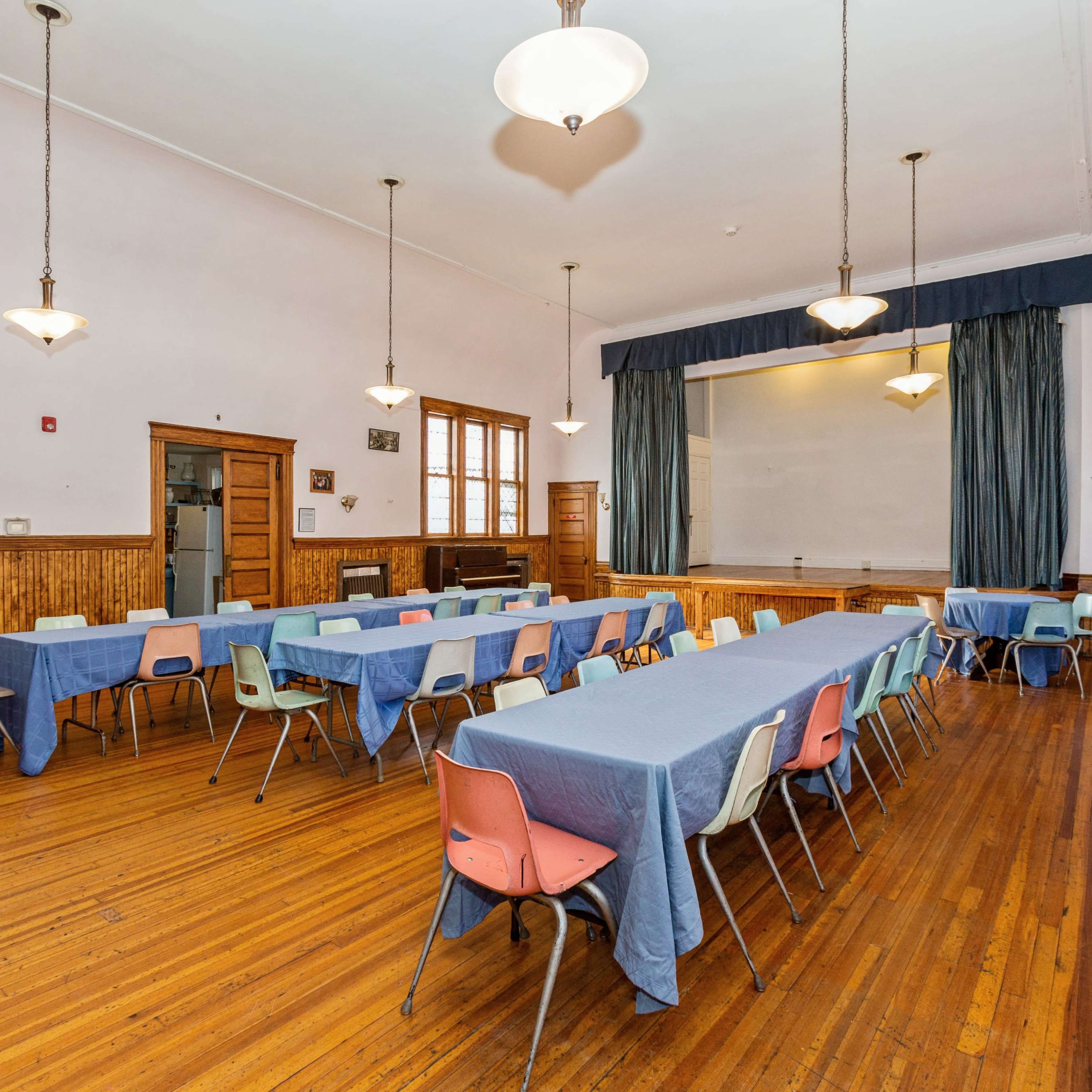 A spacious room with wooden floors, lined with several tables covered in blue cloth and arranged with colorful chairs.
