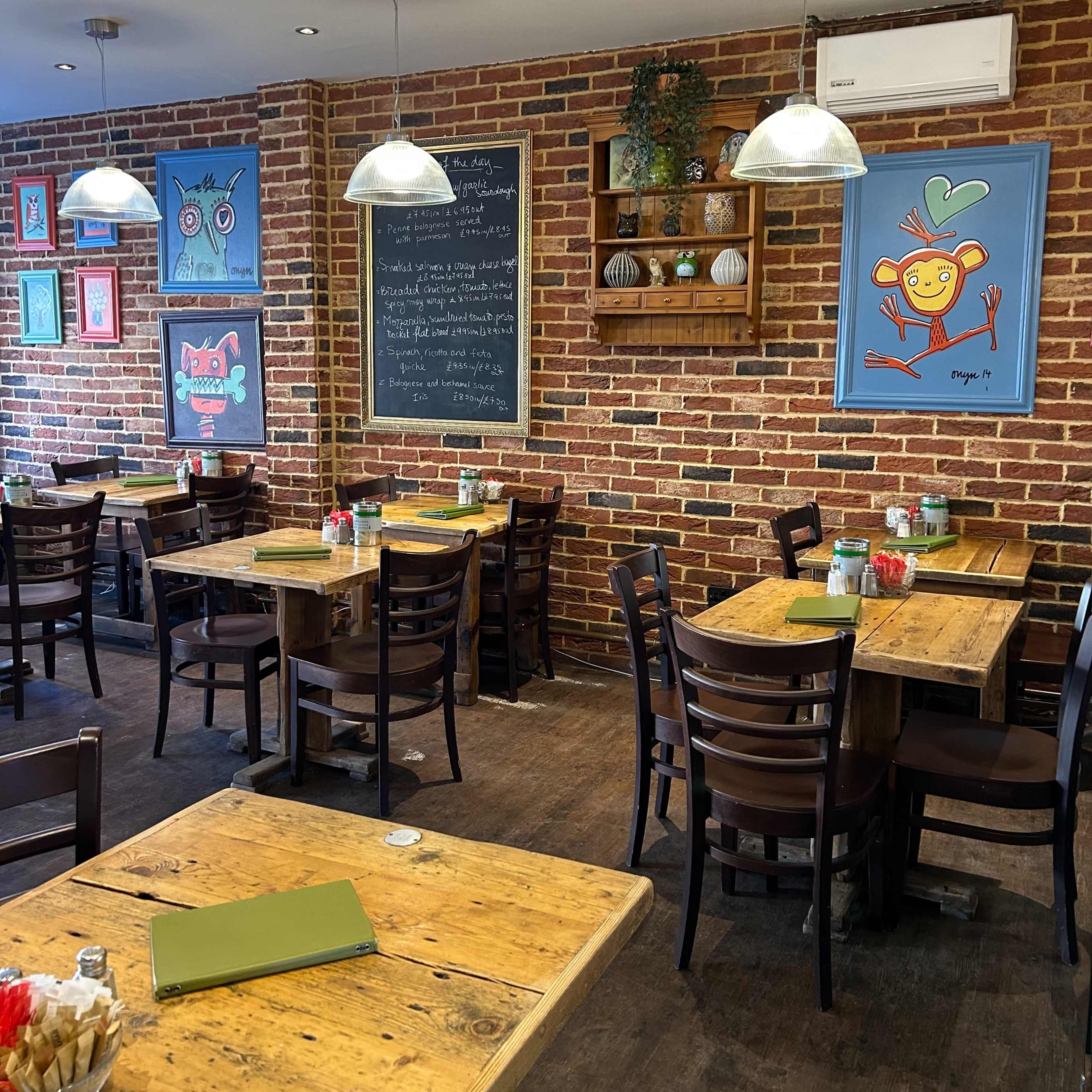 A cozy restaurant interior with wooden tables, dark chairs, and a brick wall adorned with colorful artwork and a chalkboard.